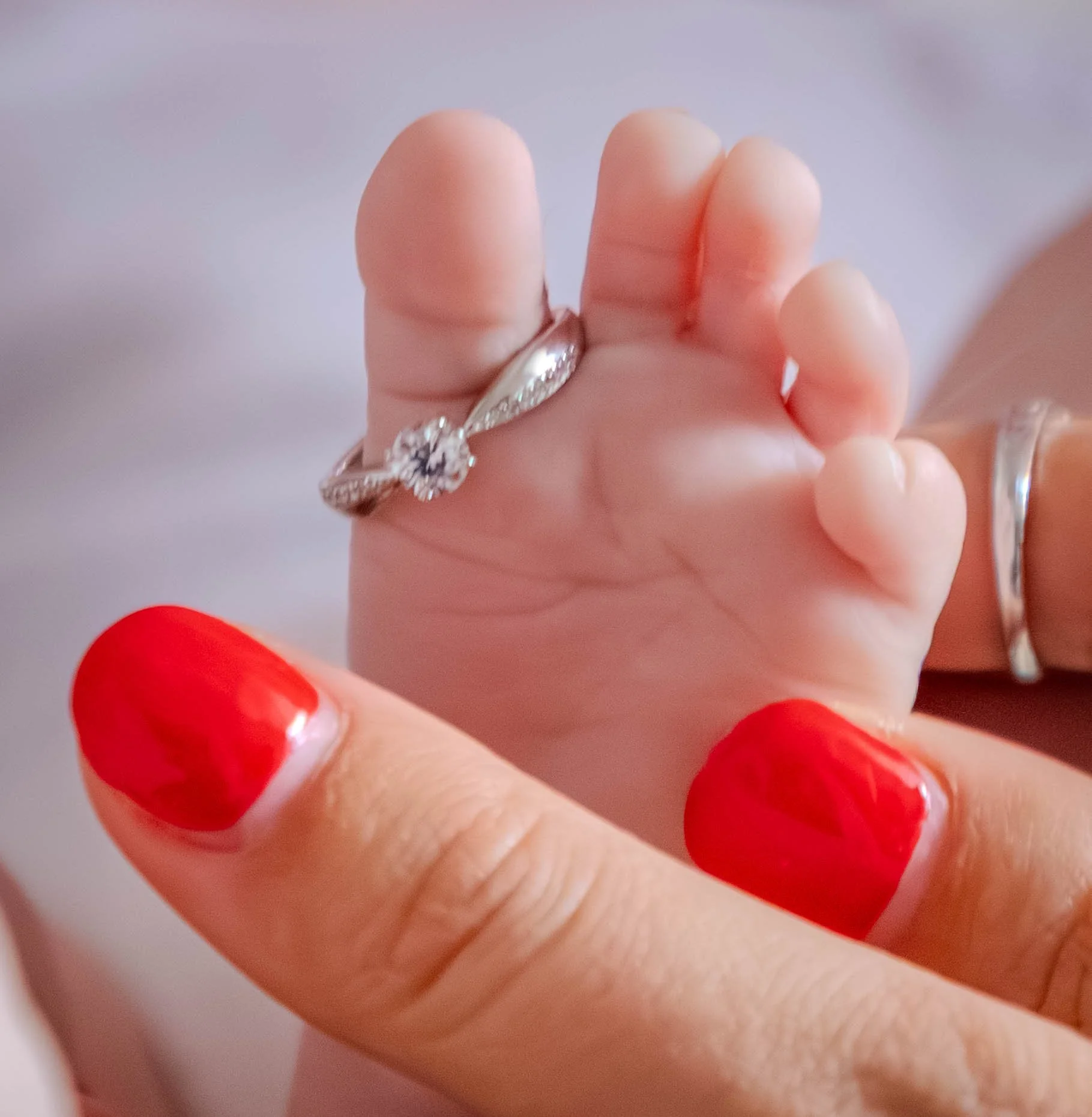Une main d'adulte avec des ongles peints en rouge tenant un petit doigt d'une main d'enfant. La main de l'enfant porte une bague en argent avec un diamant.
