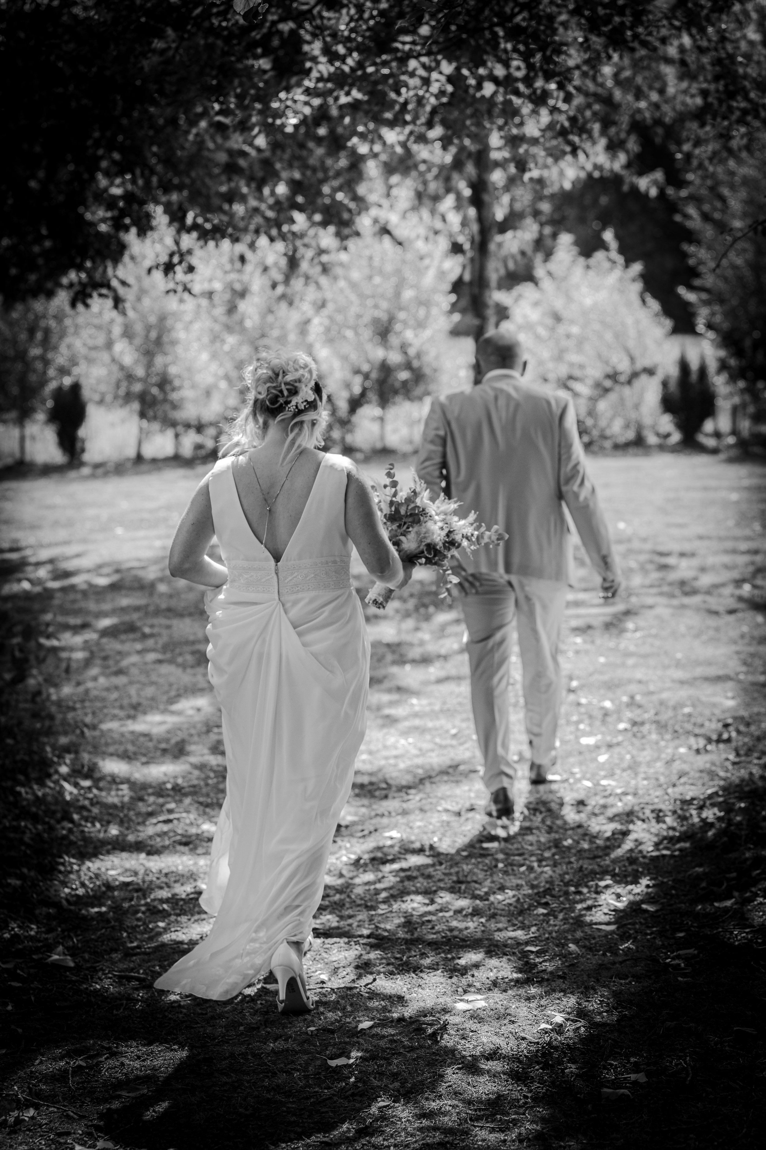Une femme en robe de mariage et un homme en costume marchent dans un parc, vue de dos, en noir et blanc.