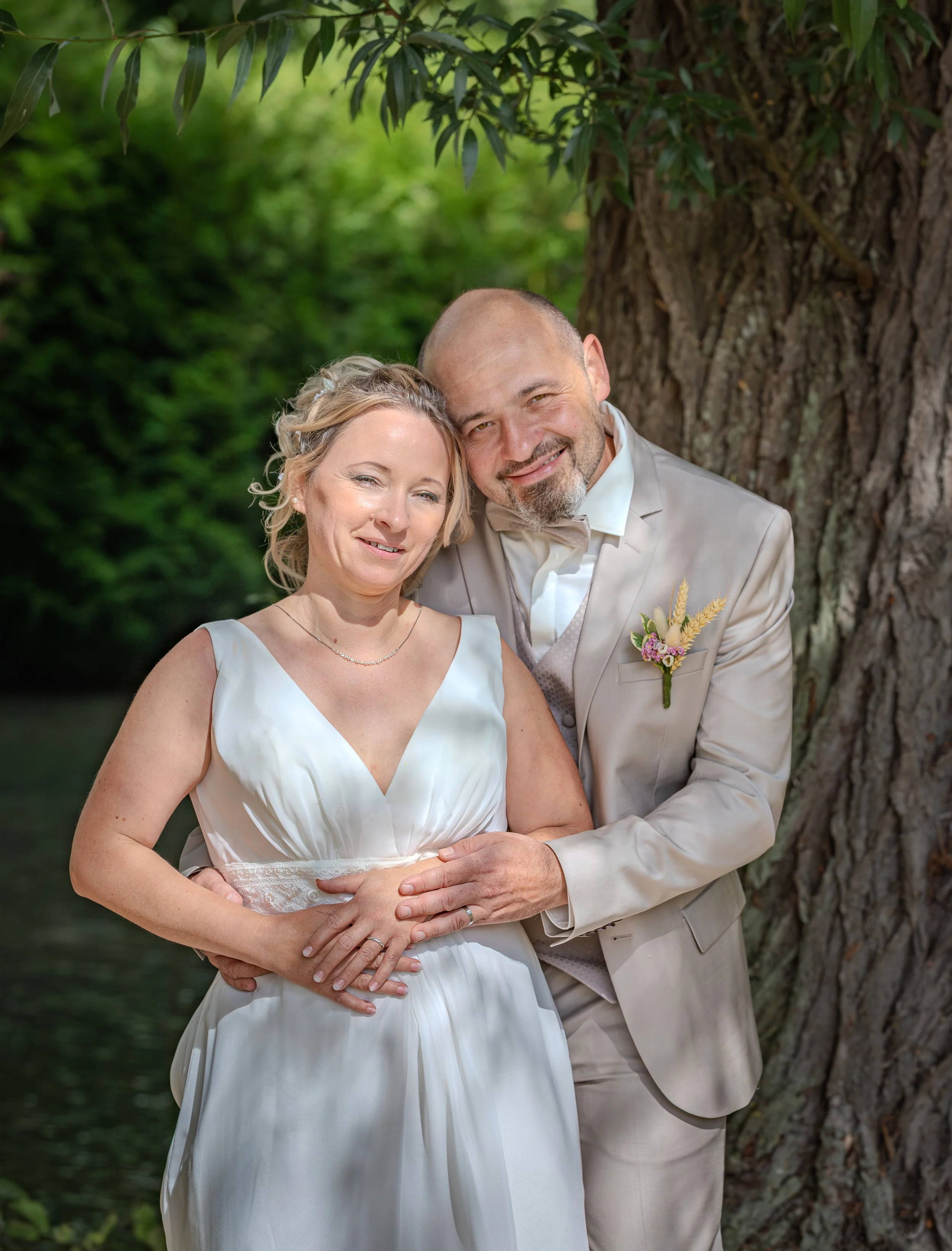 Un couple de mariés pose devant un gros arbre en plein air, dans un environnement verdoyant, durant leur mariage.