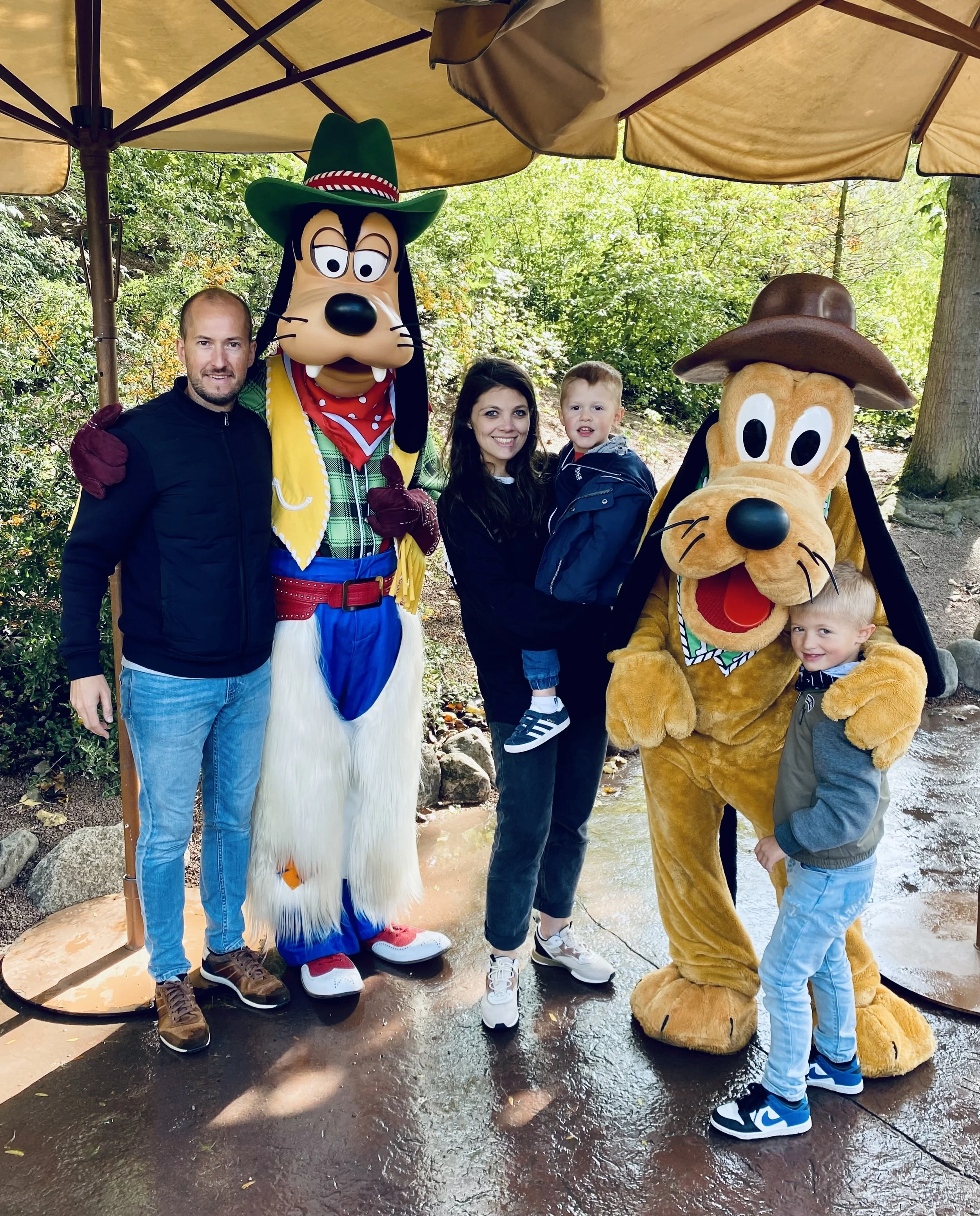 famille avec deux enfants pose avec des personnages de Disney, Pluto et Goofy, à l'extérieur sous un grand parasol dans un environnement verdoyant.