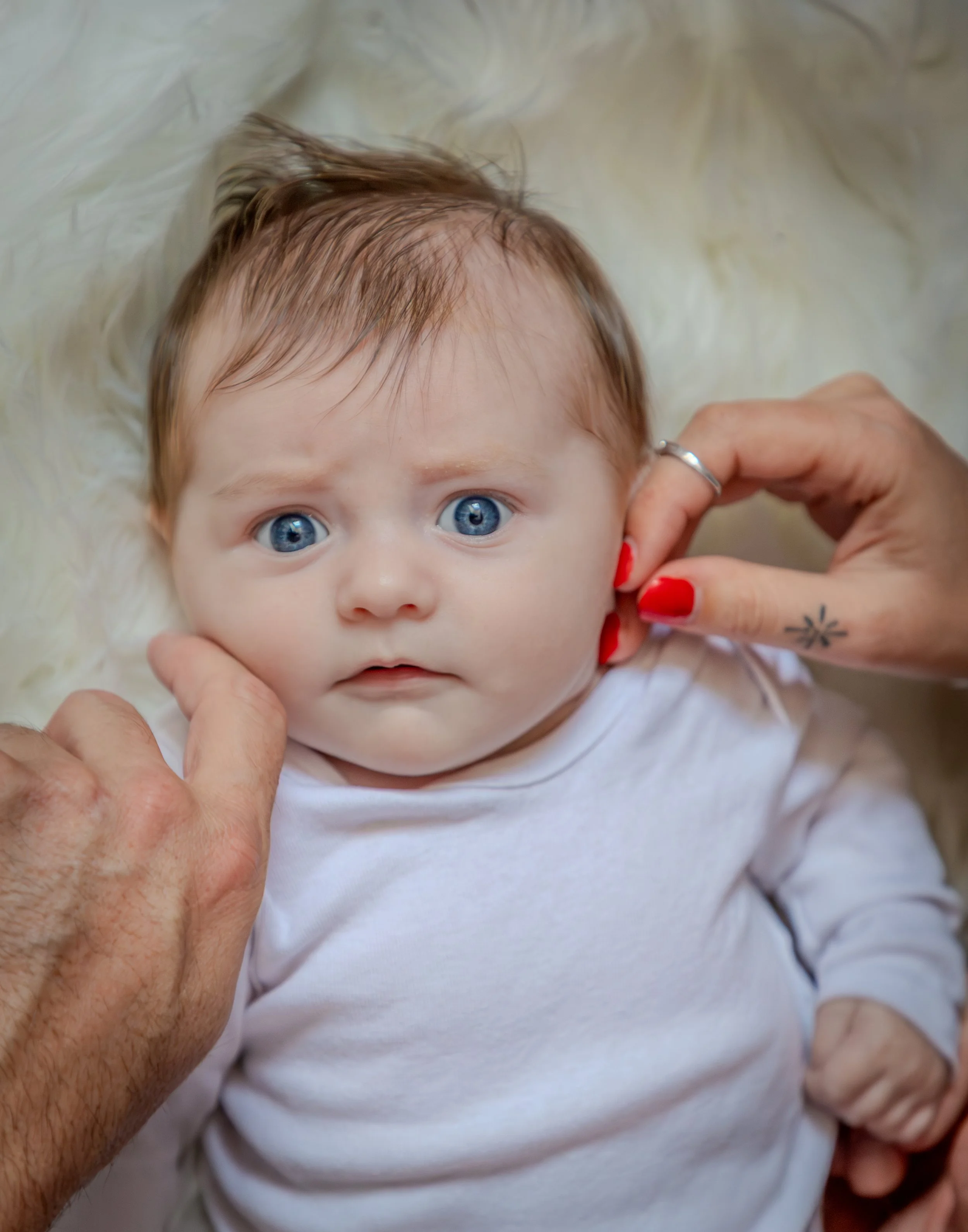 Un bébé aux yeux bleus, allongé sur une surface douce, avec une personne ajustant ses oreilles, habillé d'une chemise blanche.