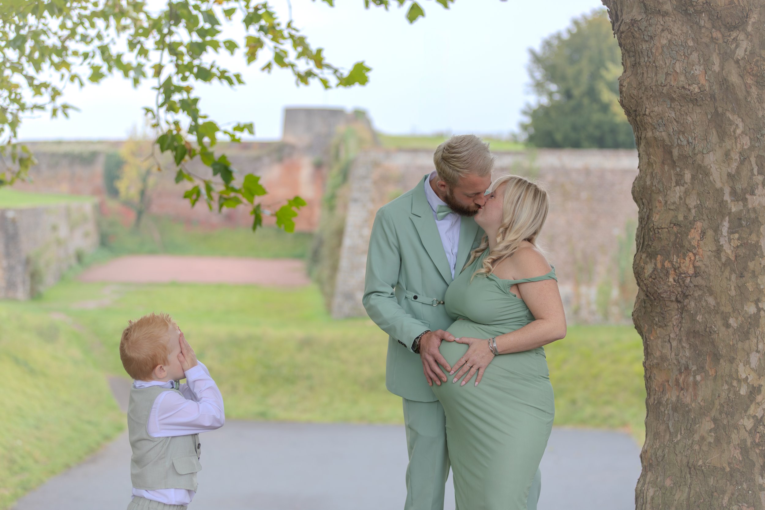 Un couple enceinte s'embrassant sous un arbre, avec un jeune garçon en veste beige qui couvre ses yeux, dans un parc avec un mur en pierres en arrière-plan.