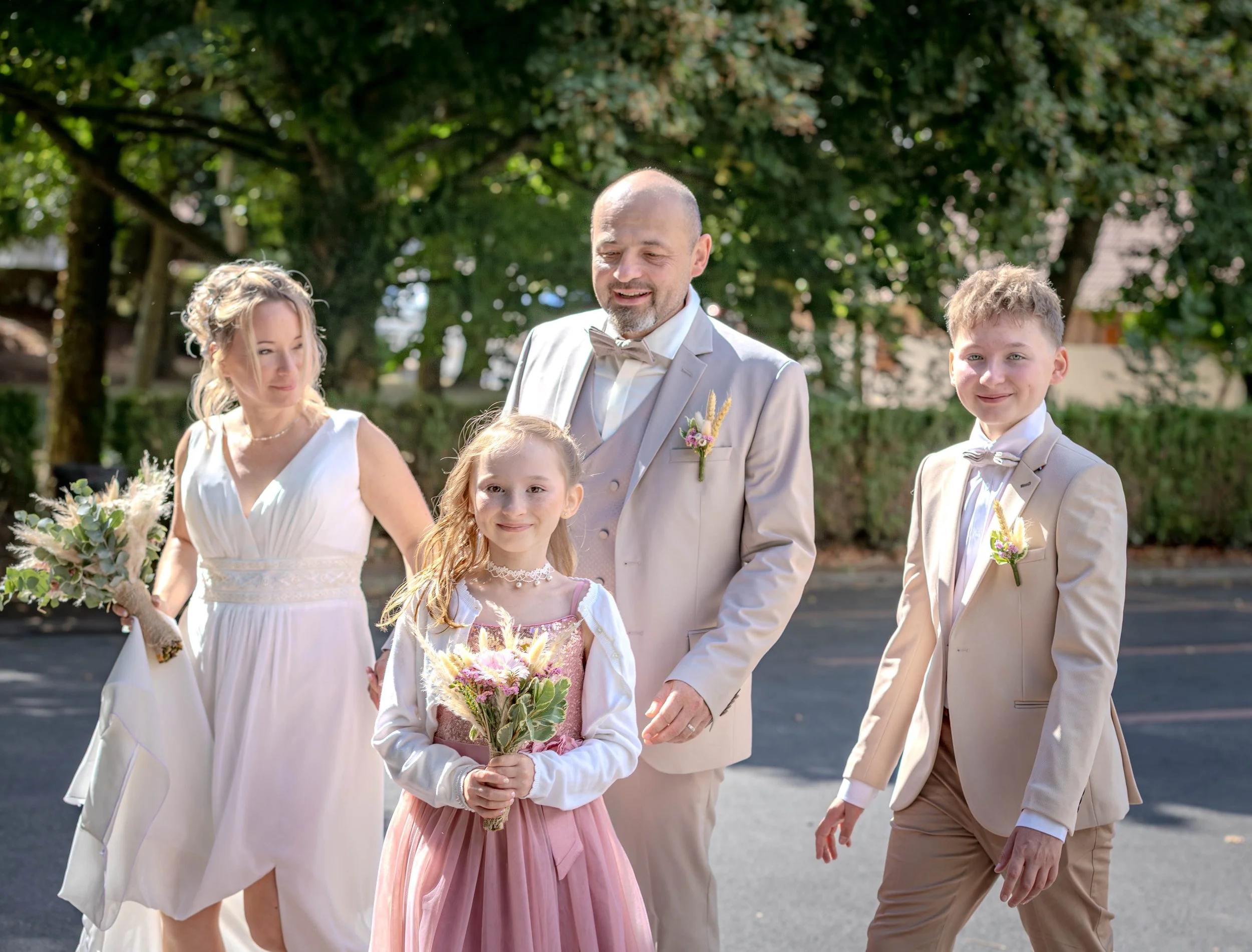 Groupe de cinq personnes habillées en vêtements de mariage, se tenant et marchant en dehors, avec des arbres en arrière-plan.