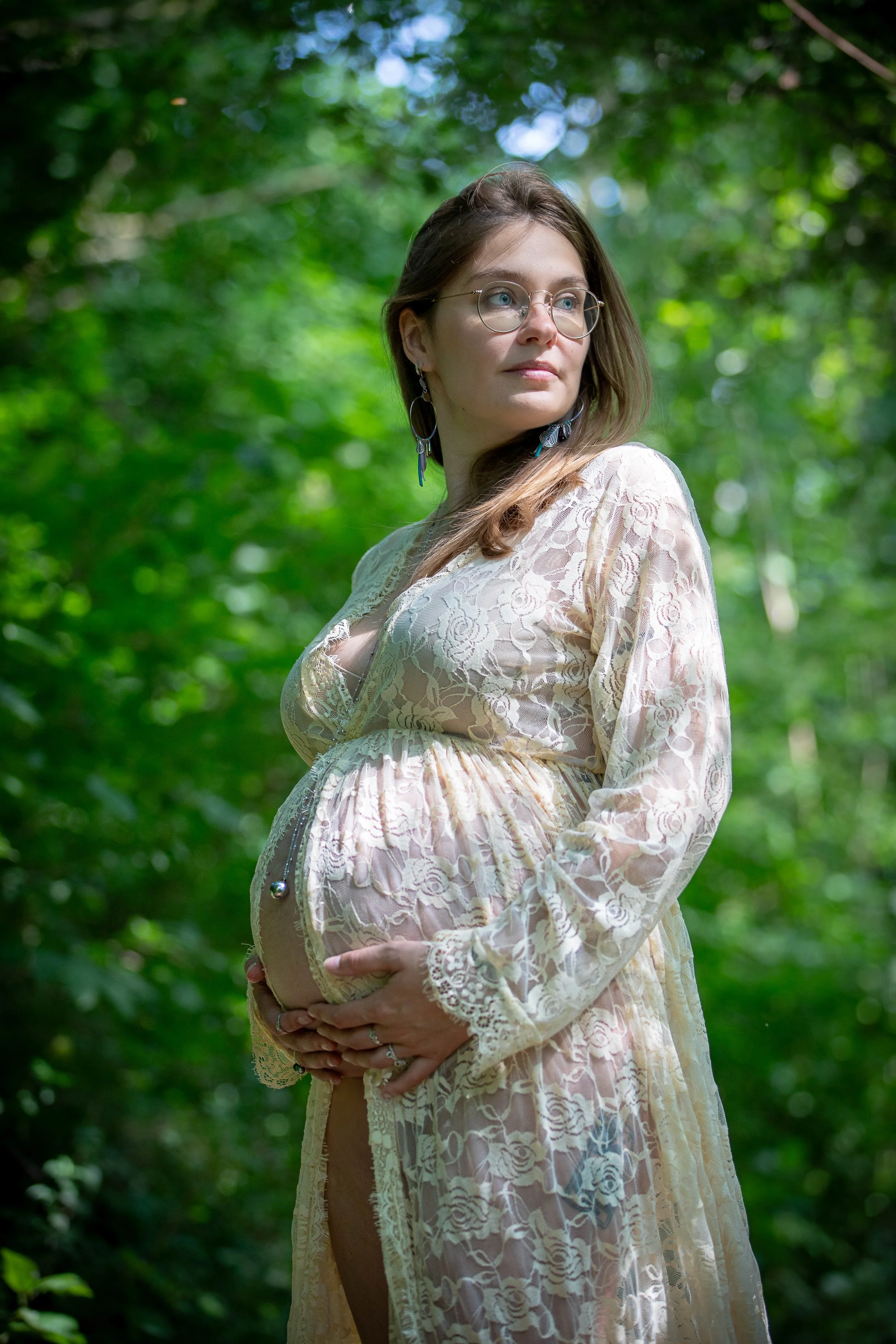 Femme enceinte portant une robe en dentelle crème, posant dans une forêt verte, tenant son ventre avec les mains, regardant vers la droite.