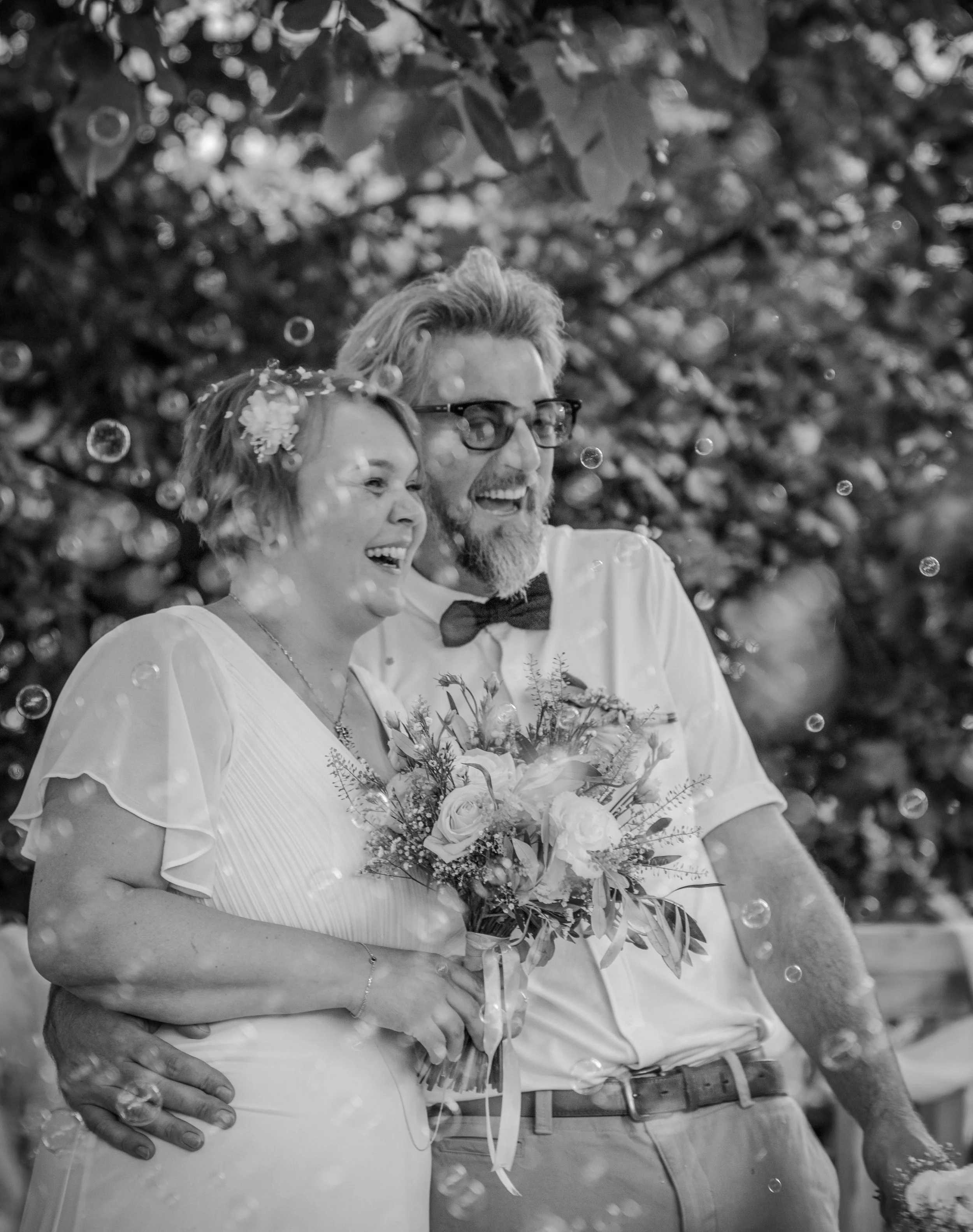 Un couple de mariage heureux posant ensemble, la femme tient un bouquet de fleurs, en noir et blanc, à l'extérieur avec des feuilles d'arbres en arrière-plan.