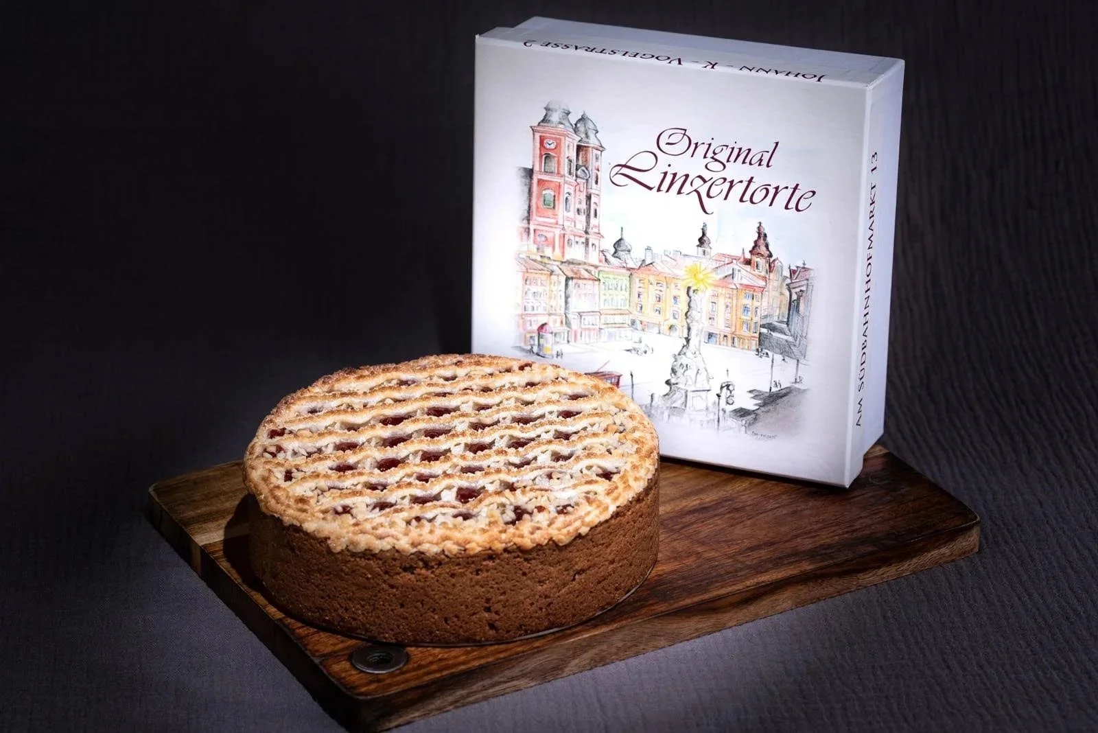 Ein runder Kuchen mit Streifenmuster auf einem Holzbrett, im Hintergrund eine Packung mit der Aufschrift "Original Linsertorte" und einer Illustration einer Stadtansicht.