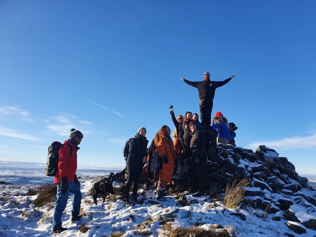 Group of hikers celebrating on a snowy hilltop with a rock cairn, one person standing on top with arms outstretched, others smiling and posing, clear blue sky in background.