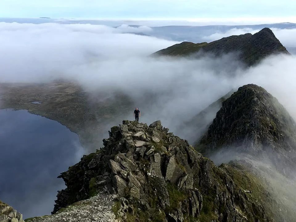 A person standing on a rocky mountain ridge with clouds and mountain peaks in the background.