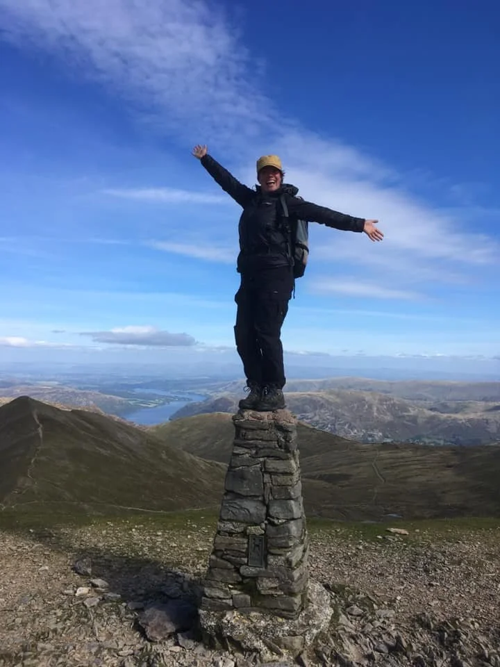 Person standing on top of a tall stone structure with arms raised in a victory pose, on a mountain with a landscape of hills and water in the background under a partly cloudy sky.