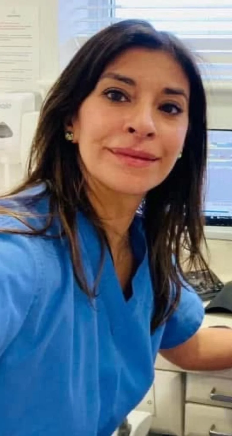 A woman with long dark hair wearing blue scrubs, sitting at a desk in front of a computer, smiling at the camera.