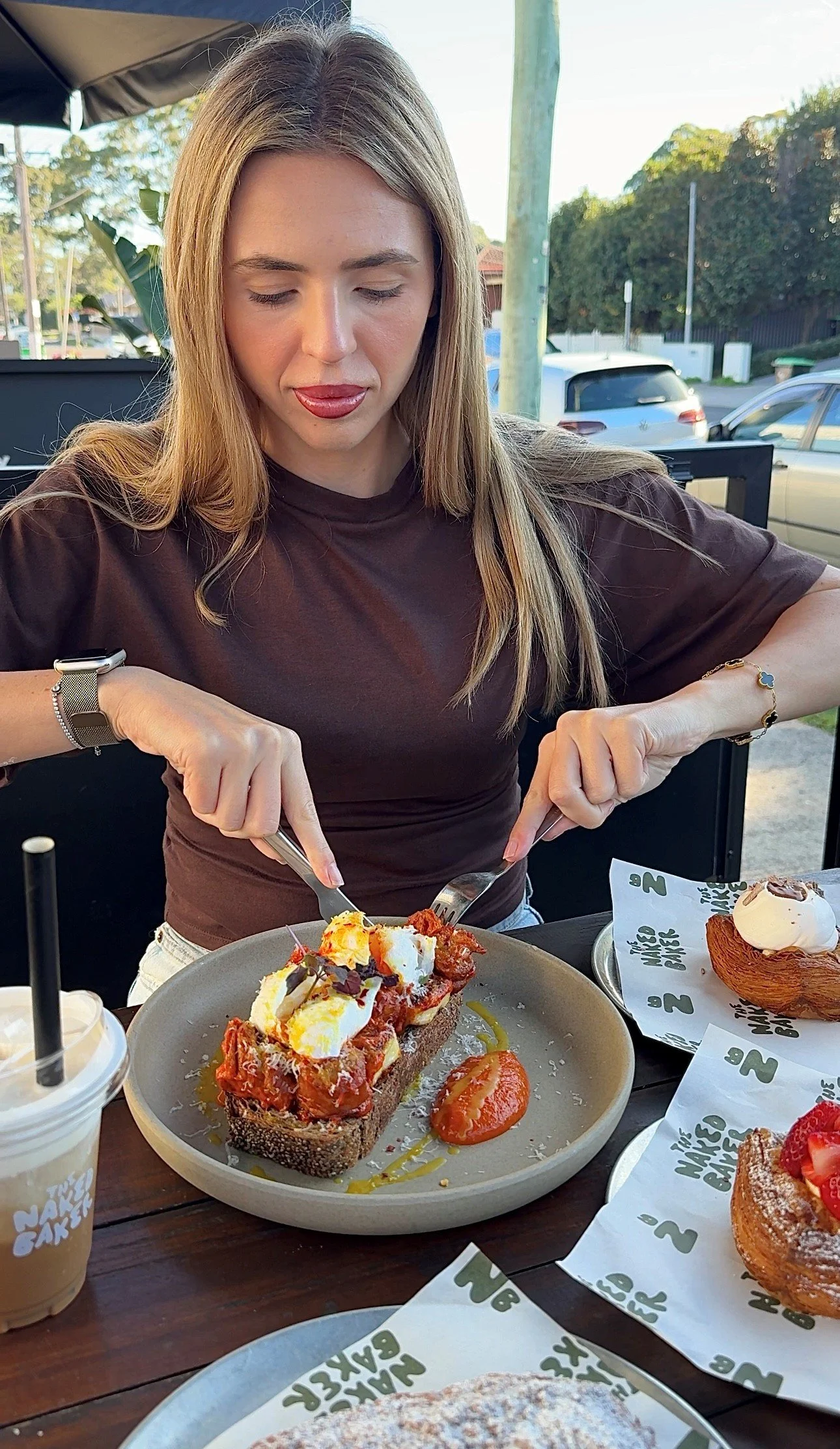 A woman sitting at an outdoor restaurant, cutting into a piece of bread topped with chopped tomatoes, cheese, and herbs. There are various desserts and a drink on the table.