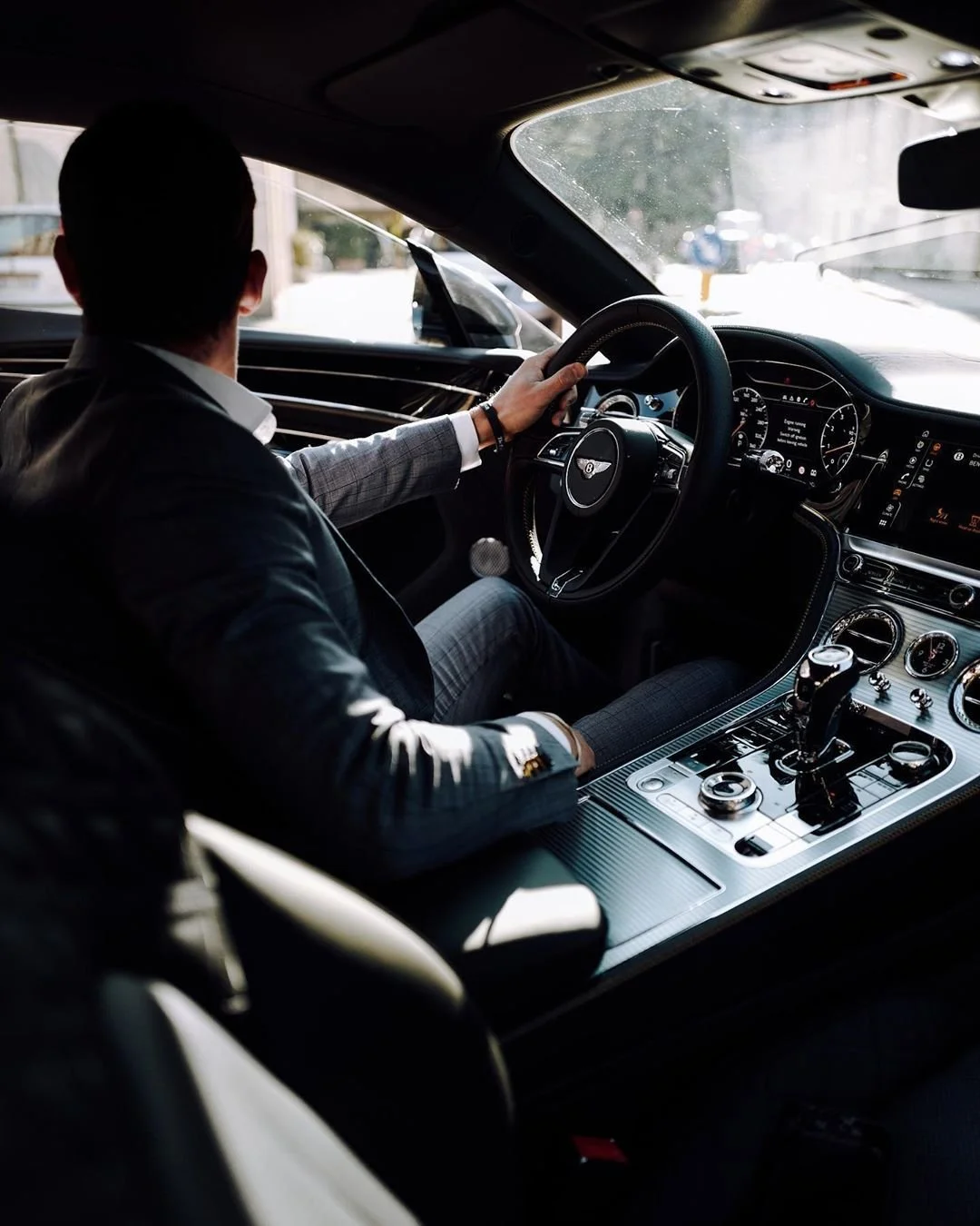 Man rijdt in een luxe auto, zichtbaar van achter en op de bestuurdersplaats, met een Garmin horloge en in een interieurlogo van Bentley.