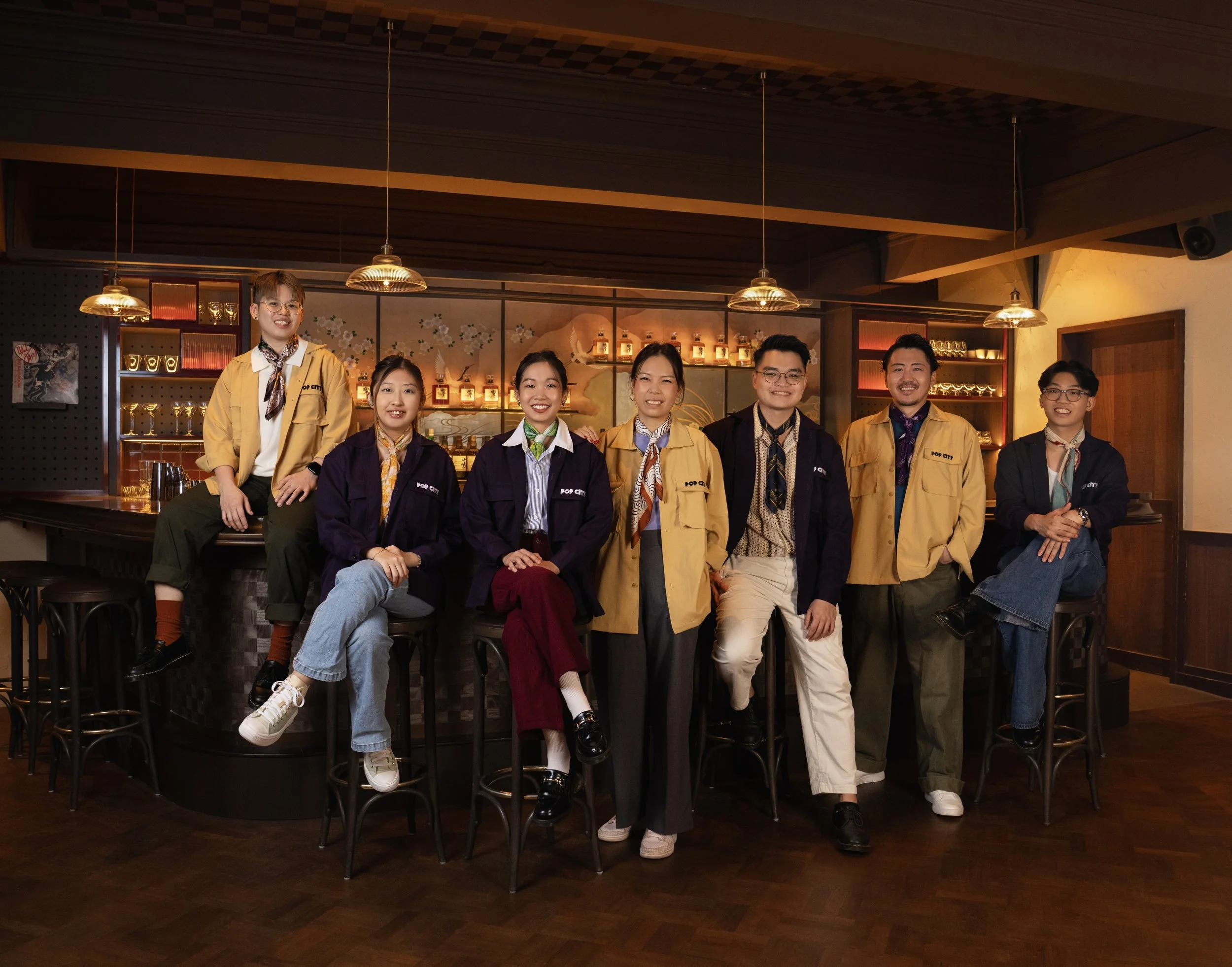 Group of nine diverse young adults smiling and posing in a cozy, dimly lit bar or café, with some sitting on bar stools and others standing behind the counter.