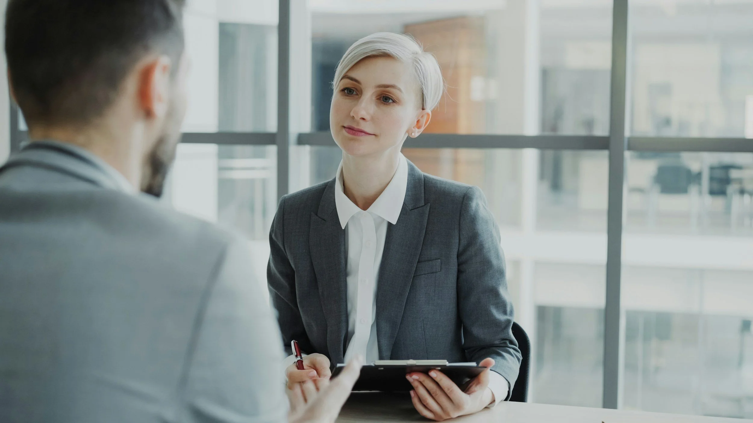 A woman in a gray business suit sitting at a table, listening attentively during a business interview or meeting with a man whose back is to the camera. She is holding a tablet or clipboard.