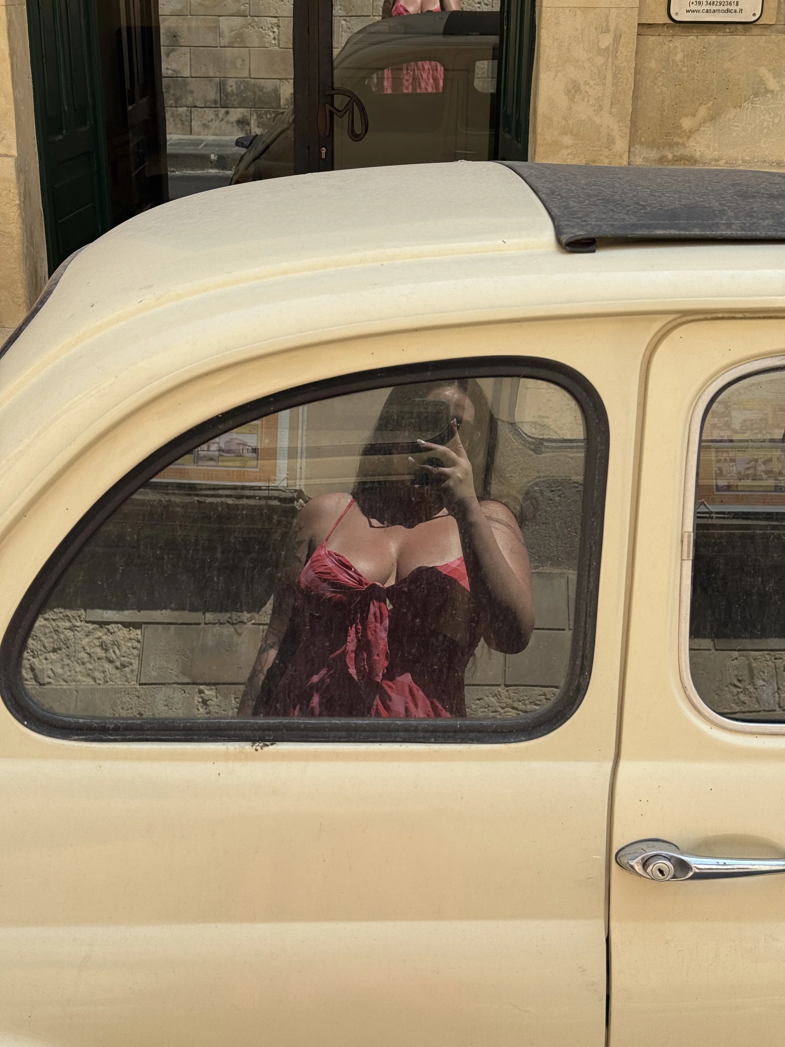 A reflection of a woman wearing a red dress is seen in the window of a vintage beige car.