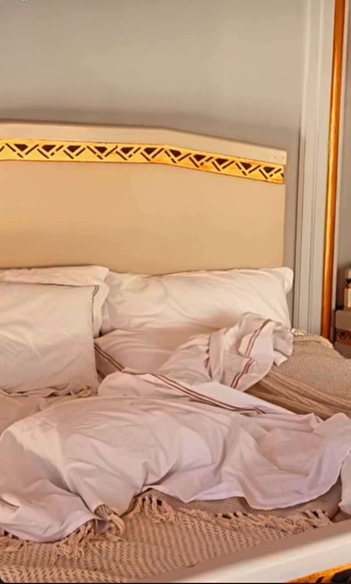 An unmade bed with disheveled white sheets and pillows, located in a room with a headboard that has a yellow decorative strip with black geometric patterns.