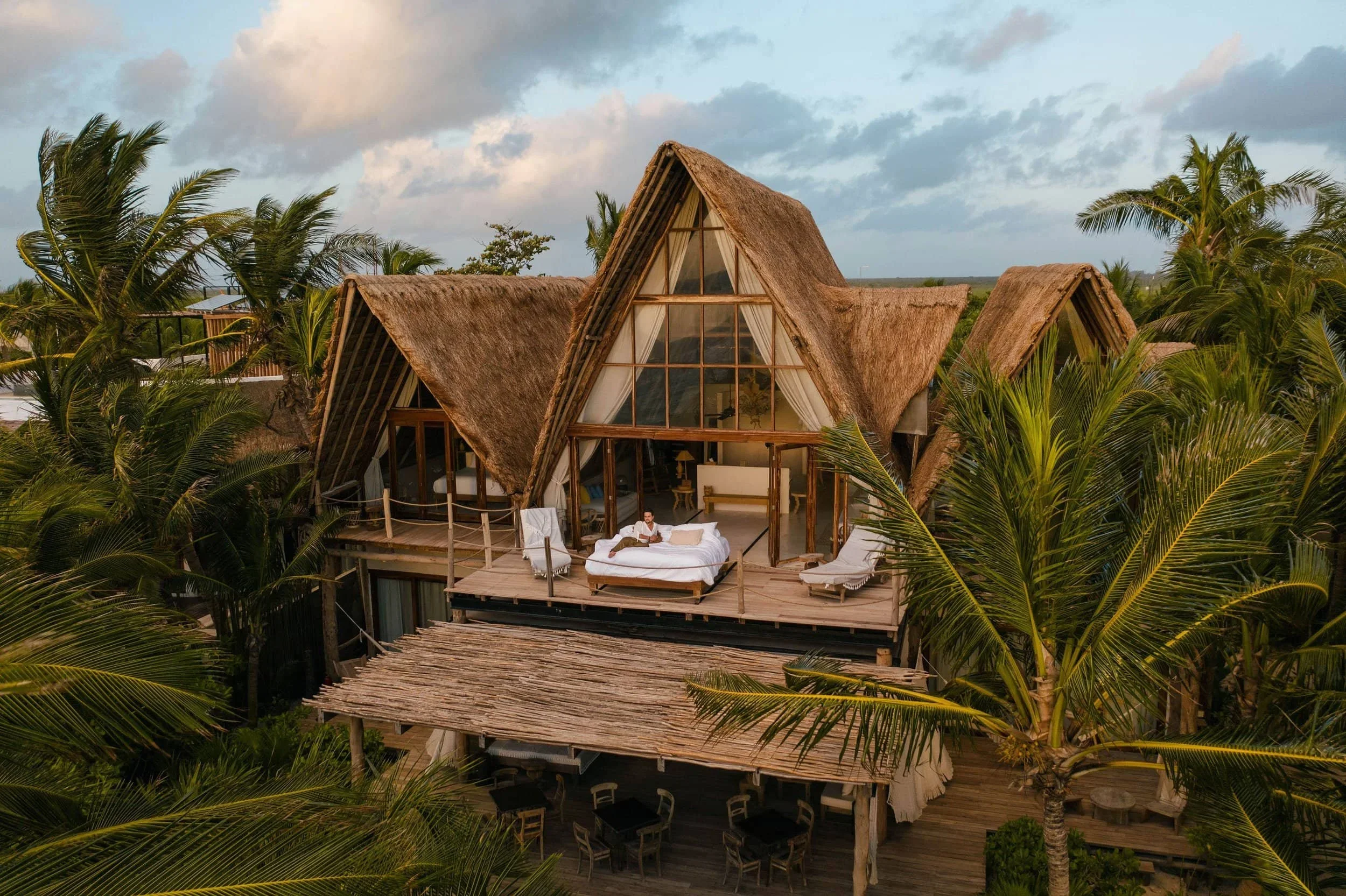 A tropical resort with thatched roof buildings, palm trees, and a wooden deck with outdoor furniture. A person is sitting on a bed on the deck, surrounded by lush greenery.