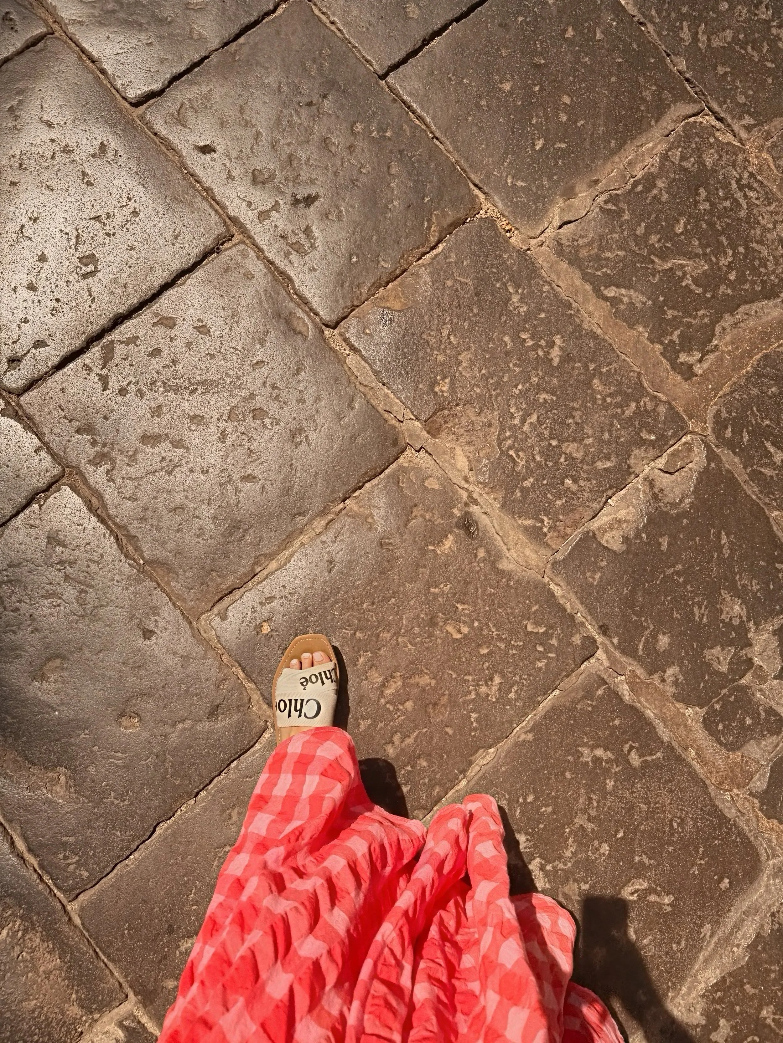 A person's feet wearing Chloé brand sandals, standing on a dark stone tiled floor. The person is dressed in red and white checkered pants.
