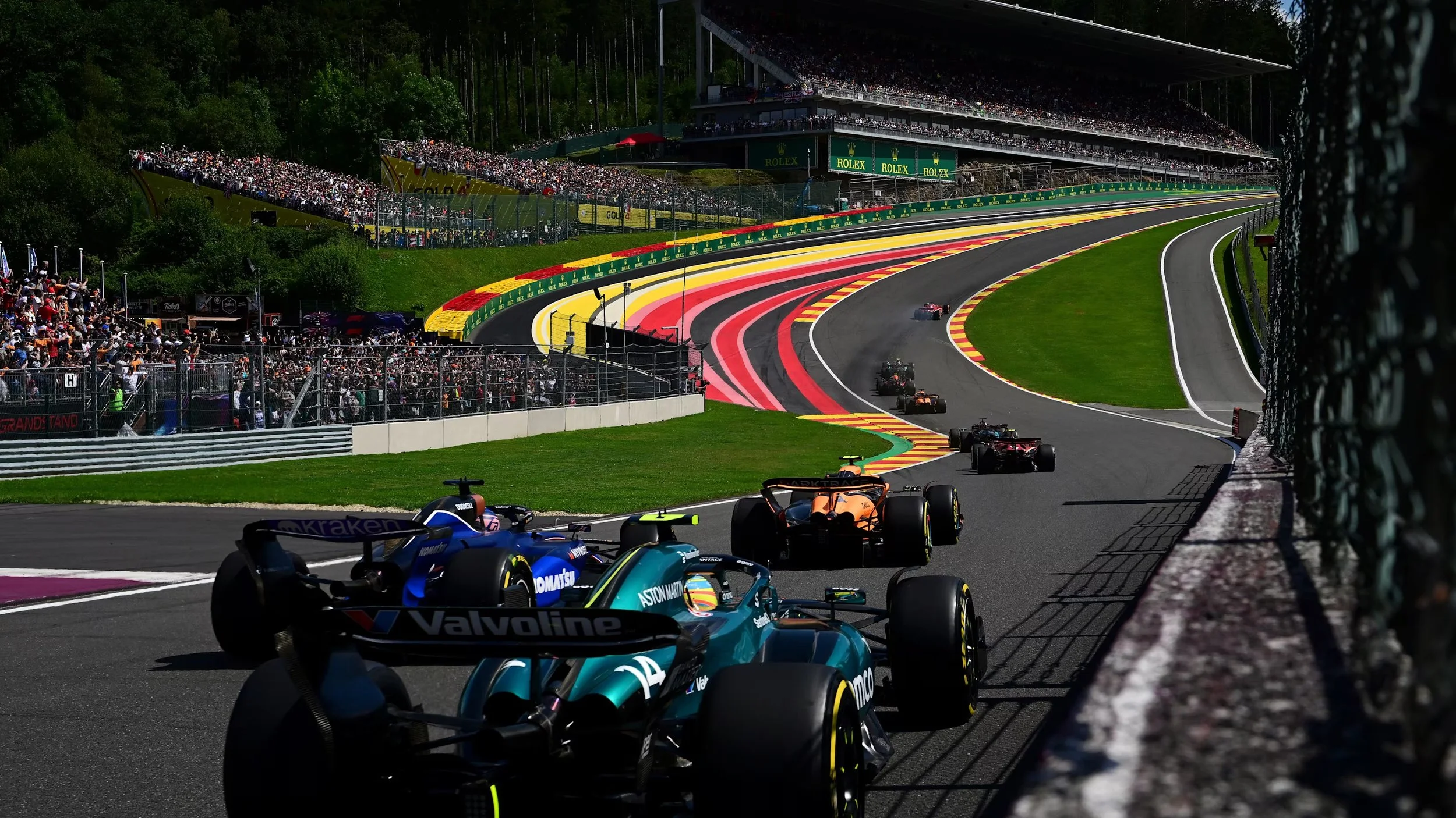 Formula 1 cars racing on a track with spectators in the stands and greenery around, viewed from the side of the track.