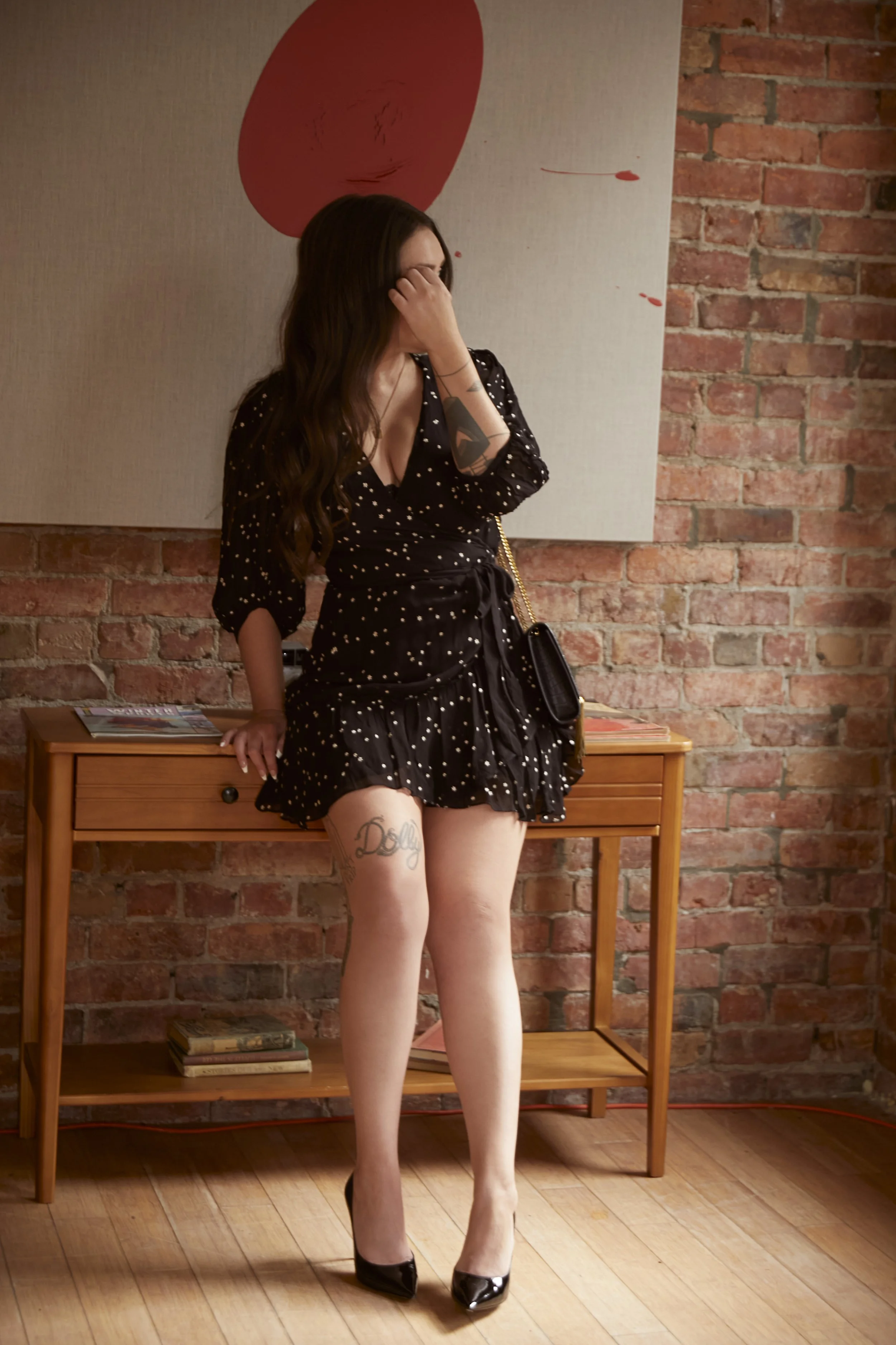 A woman with long wavy brown hair wearing a black dress with white polka dots, black high heels, and a gold chain strap purse, standing in front of a brick wall and wooden furniture.