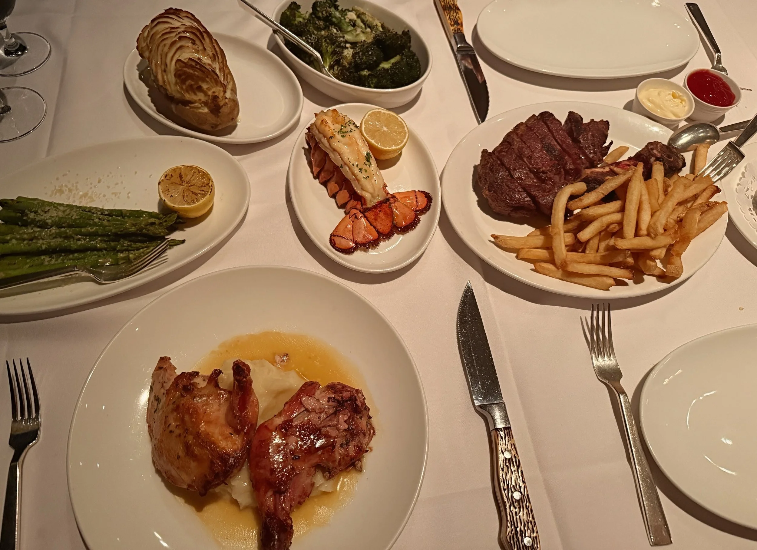 A dinner table set with various dishes, including asparagus, broccoli, fish, steak, French fries, and bread, with utensils and condiments.
