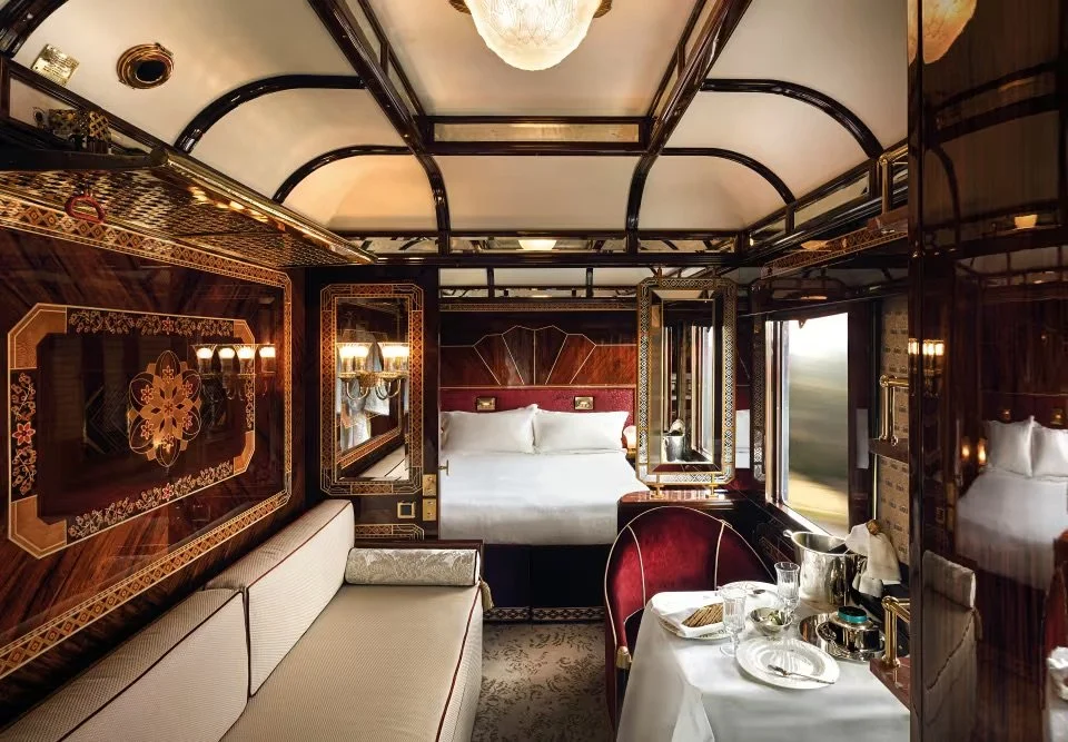 Luxurious train cabin with ornate woodwork, a bed with white linens, a small dining area with a table set for a meal, and elegant decor.