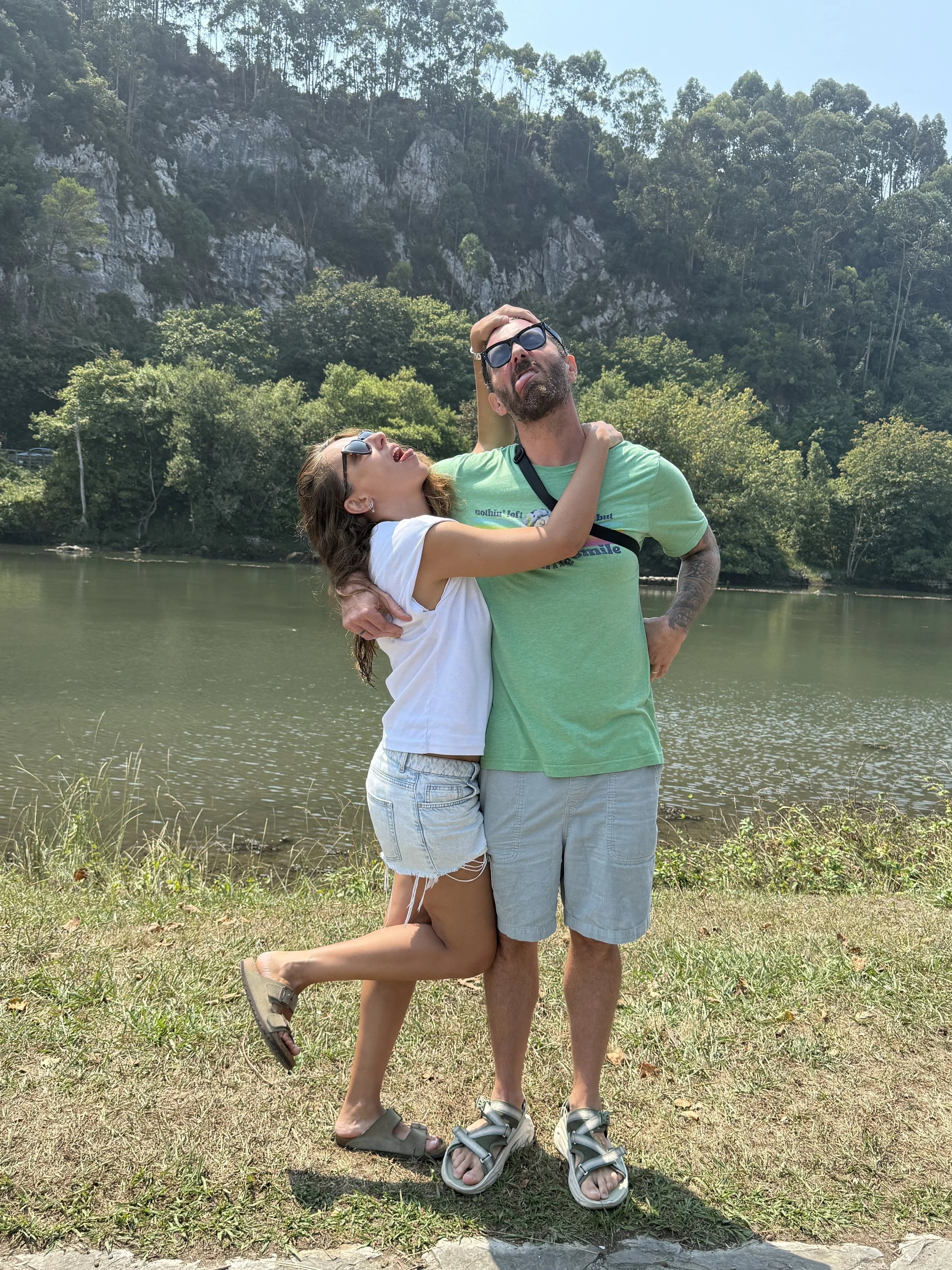 A woman and a man embrace near a body of water with trees and hills in the background. The woman is hugging the man around his neck, and the man has his arm around her waist. They are both wearing sunglasses and casual summer clothes, with the woman in a white shirt and denim shorts, and the man in a green t-shirt, gray shorts, and sandals.