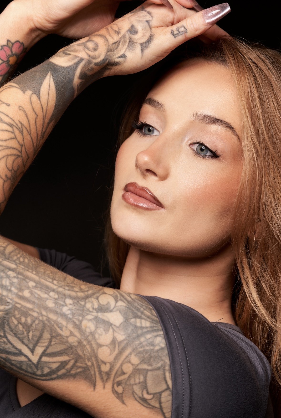 A woman with red hair and tattoos on her arm and hand poses against a black background.