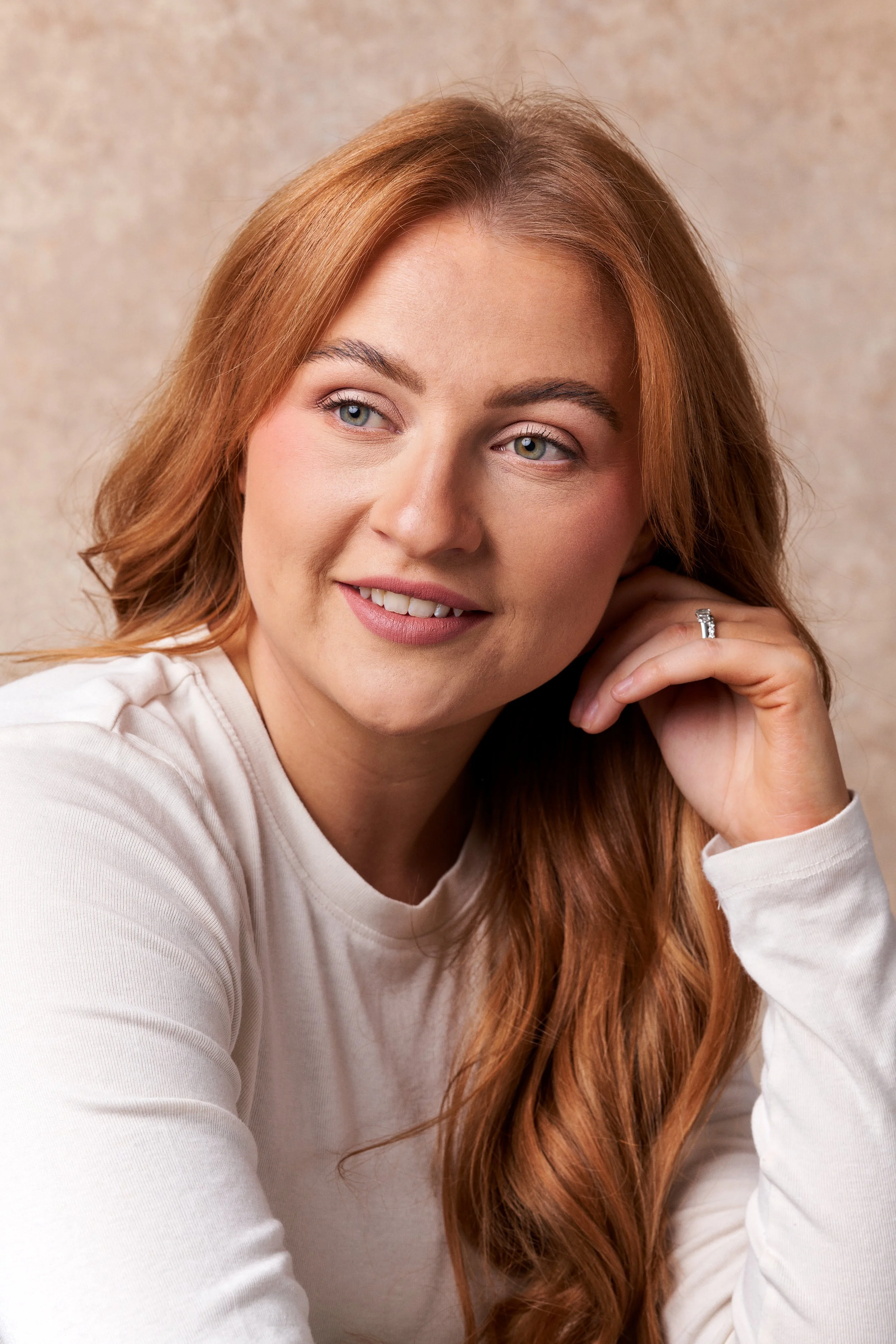 A young woman with red hair and blue eyes, smiling softly and resting her head on her hand, wearing a white long-sleeve top.