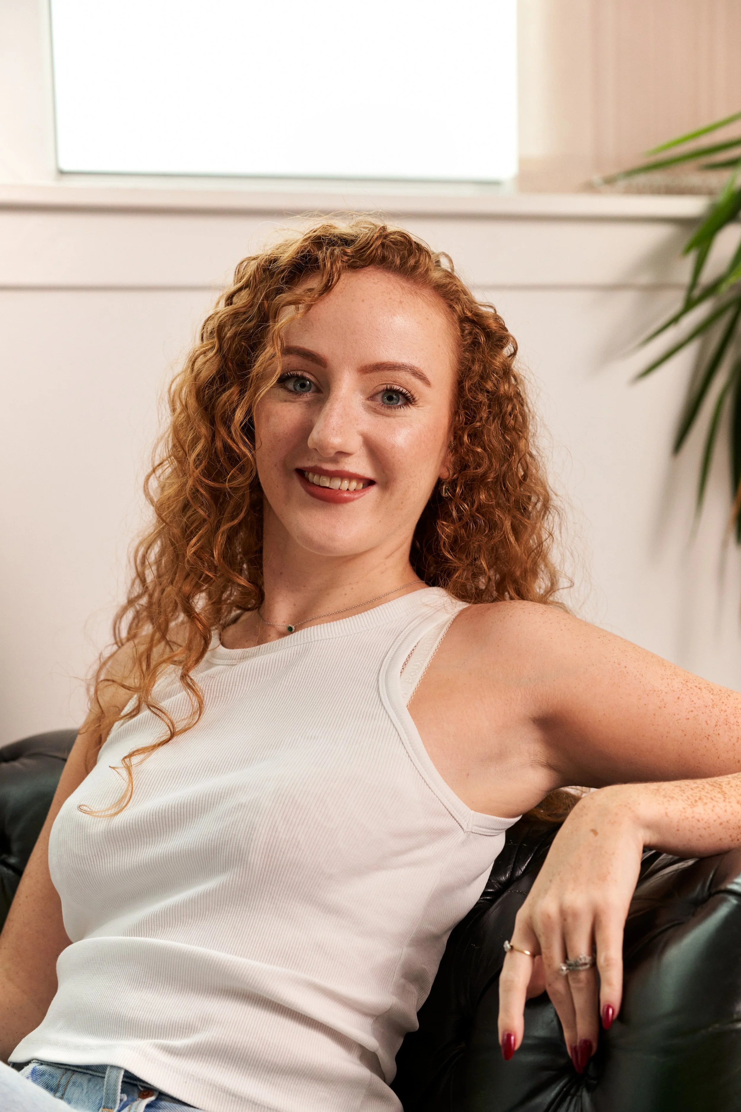 A young woman with curly red hair smiling while sitting on a black leather couch in a bright room with a plant and a window in the background.