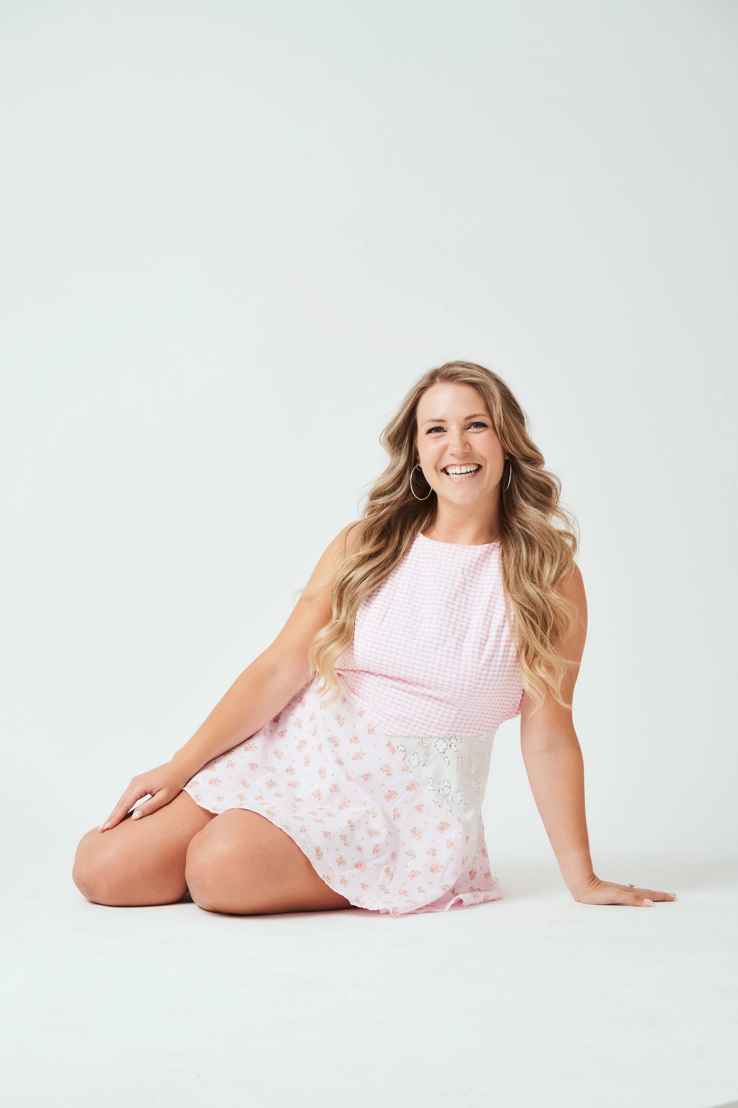 A woman with long, wavy blonde hair smiling and sitting on the floor against a plain white background, wearing a pink and white dress with lace details and hoop earrings.