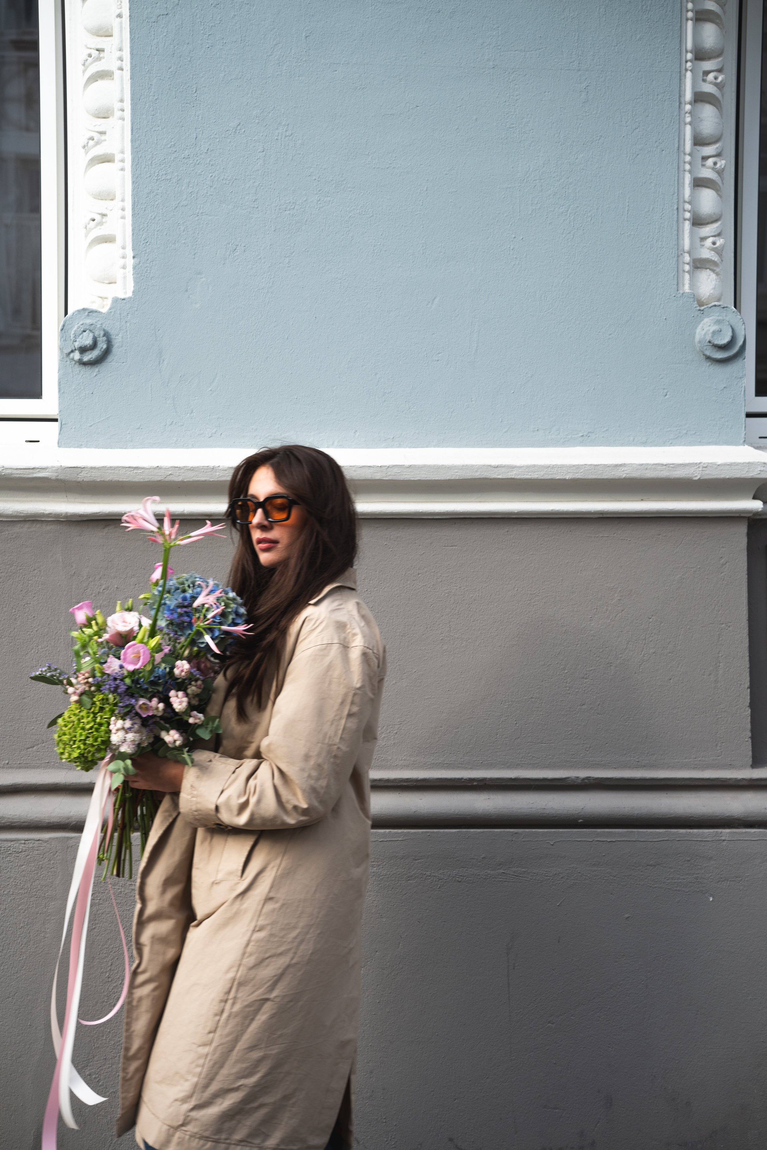 Eine Frau mit dunklen Haaren, Sonnenbrille und beigem Mantel hält einen Blumenstrauß mit bunten Blumen vor einer hellgrauen Wand.