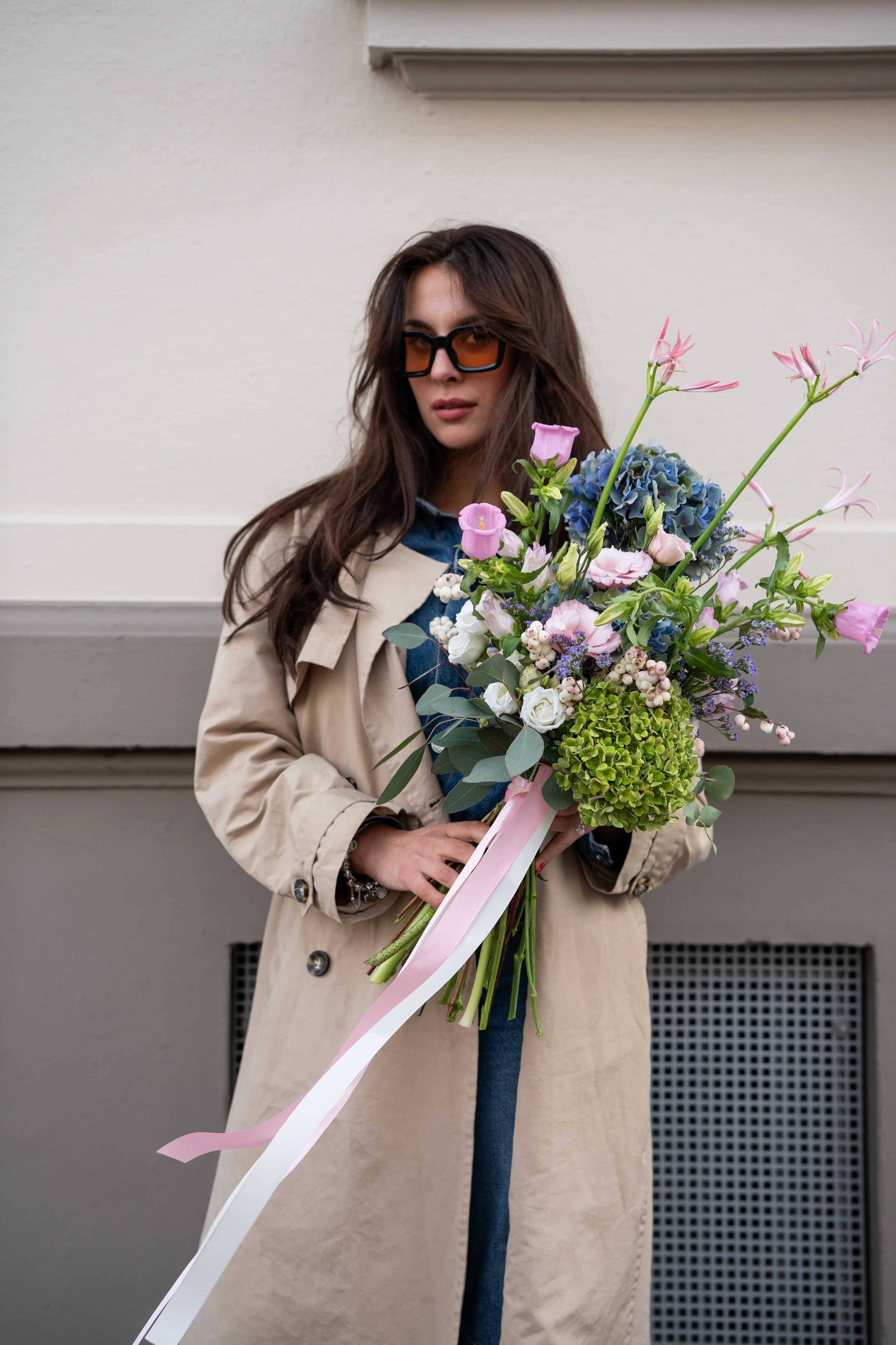 Frau mit Sonnenbrille hält einen großen bunten Blumenstrauß vor einem hellen Gebäude.