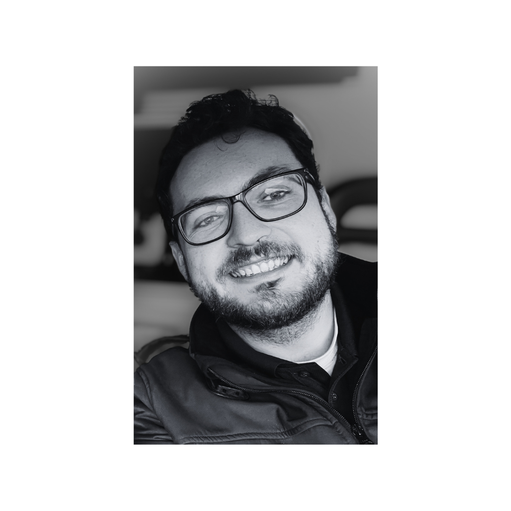 A smiling man with glasses, a beard, and dark hair, wearing a jacket and a shirt, in a close-up black and white photograph.