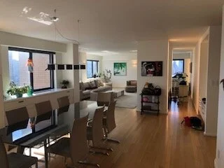 A spacious living and dining area with large windows, a dining table with chairs, a sofa, and a small cabinet, with natural light coming in.