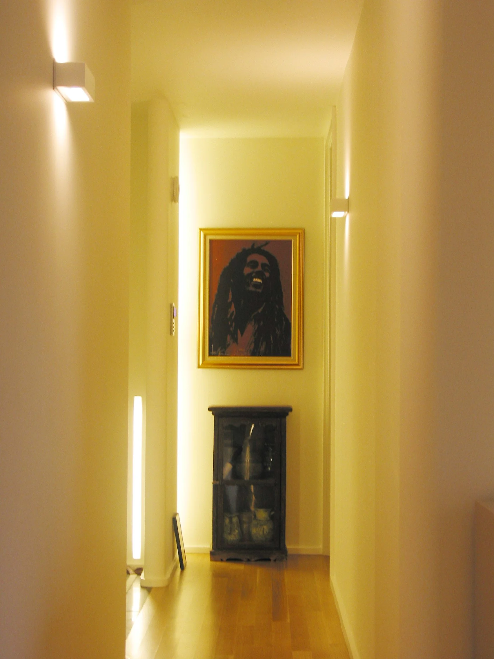 A hallway with a yellow wall, a framed picture of Bob Marley, a black display cabinet, and a floor made of hardwood.