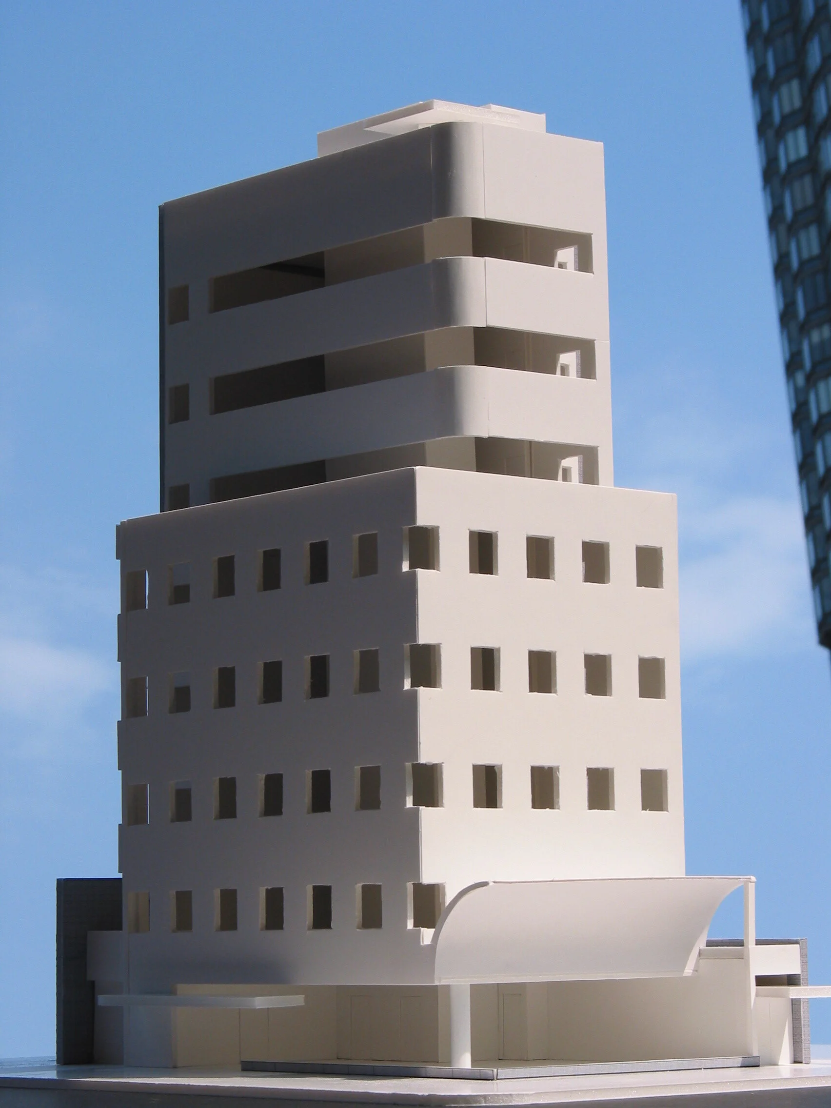 Architectural model of a modern white building with multiple floors, large rectangular and square windows, and curved design elements against a blue sky.