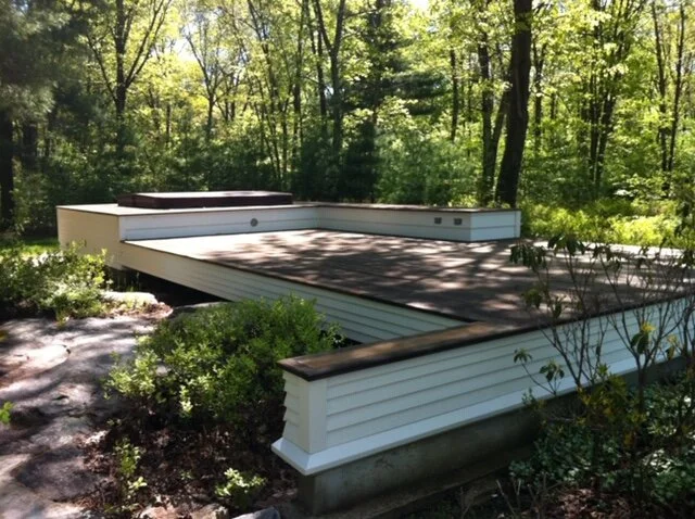 A backyard deck with a raised hot tub, surrounded by green trees and bushes, in a wooded area.