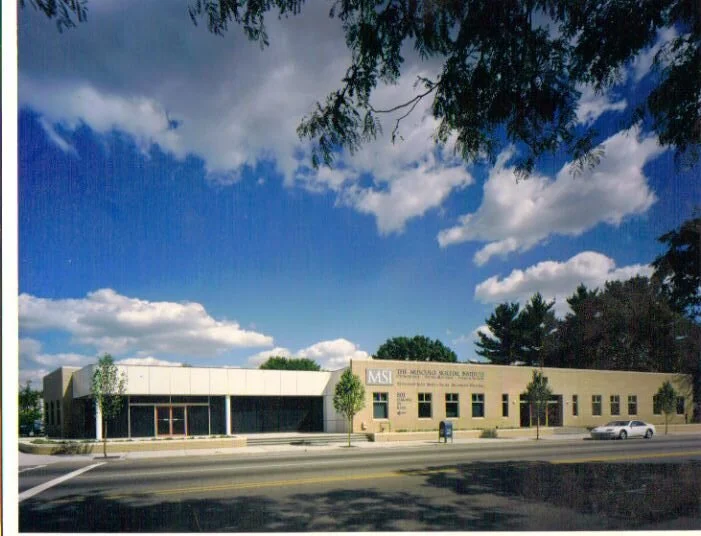 A modern school building under a partly cloudy sky, with trees in front and a parked car on the street.