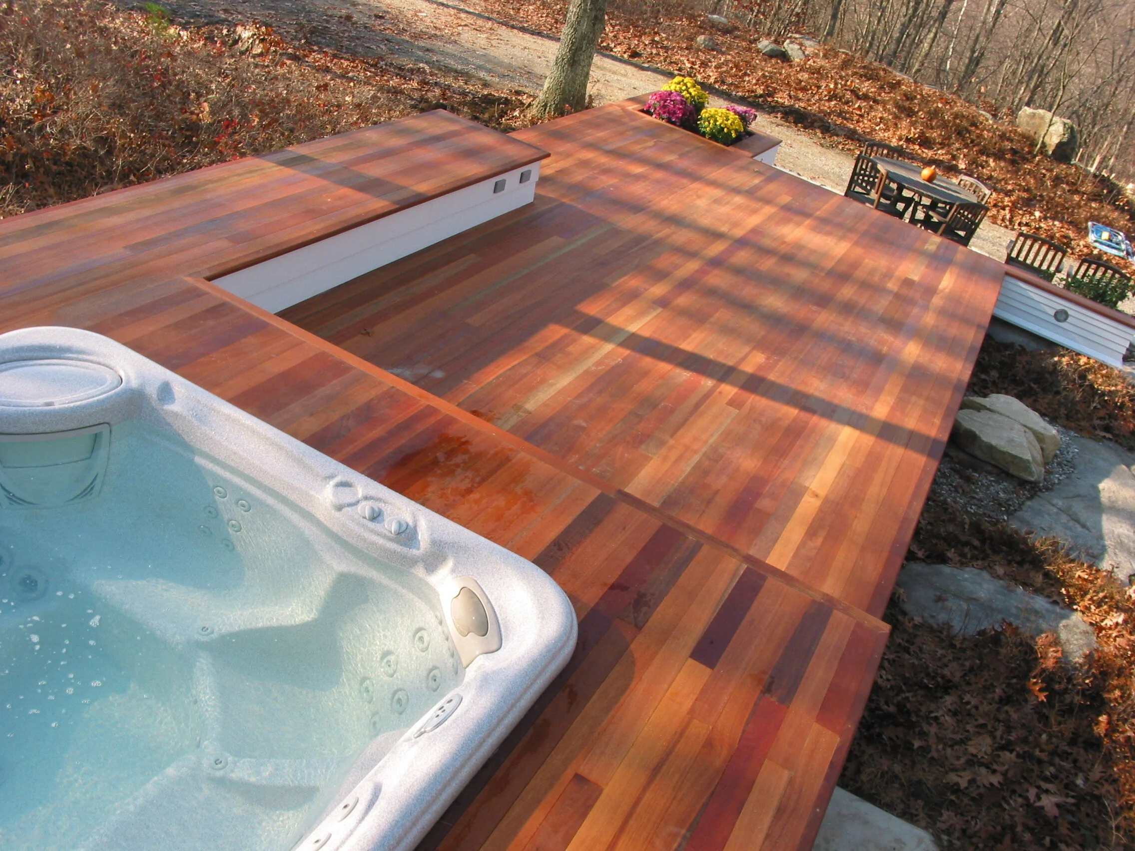 A deck with a hot tub in the foreground, wooden flooring, a dining table with chairs, and autumn leaves on the ground with trees in the background.