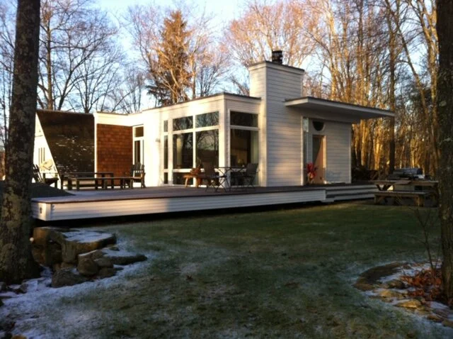 A modern house with large windows, a deck, and outdoor seating, surrounded by trees.
