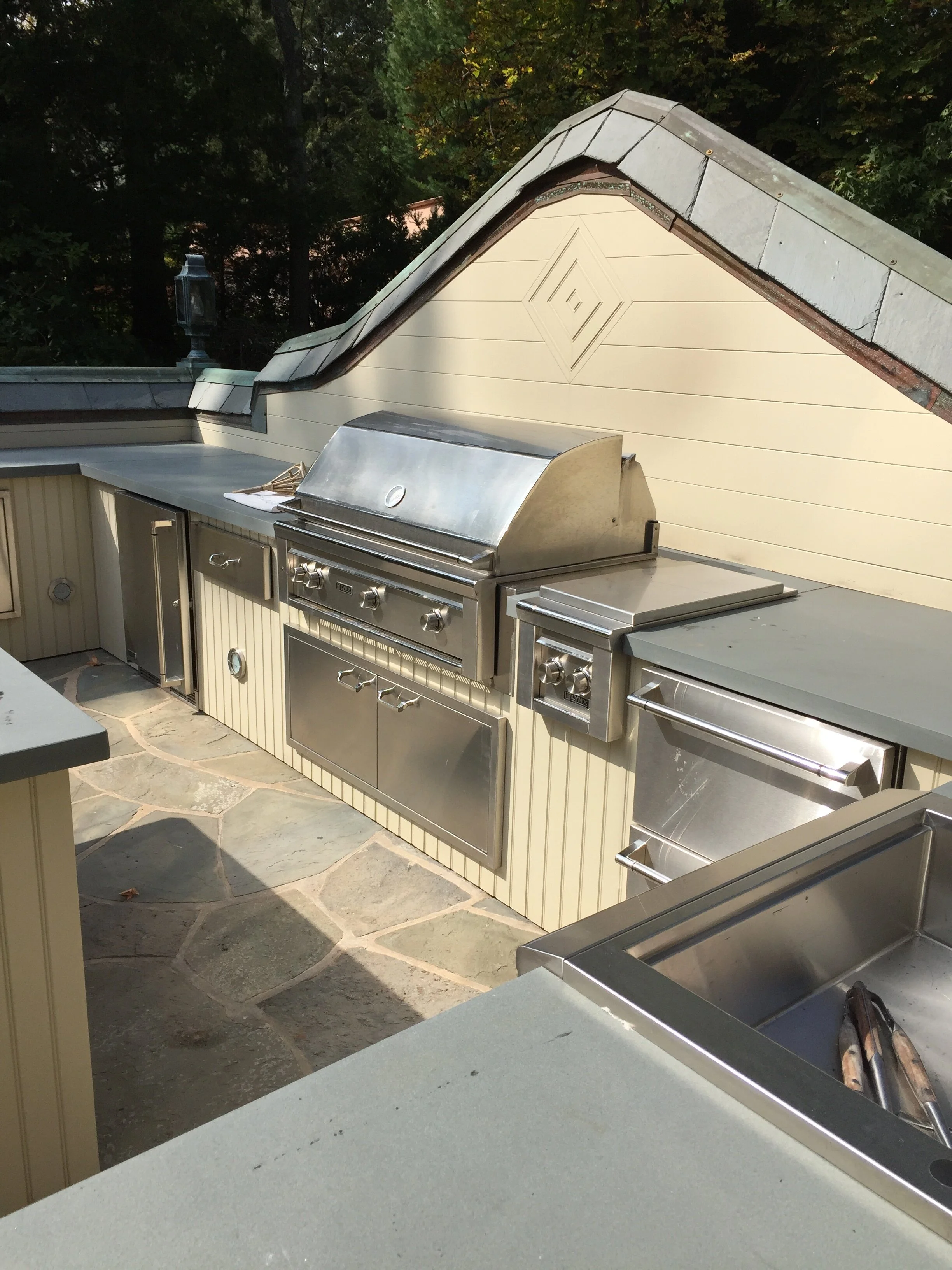 Outdoor kitchen with stainless steel appliances, including a grill, countertop, and storage drawers, set against a cream-colored wall with siding and a decorative diamond pattern, surrounded by trees and a stone patio.