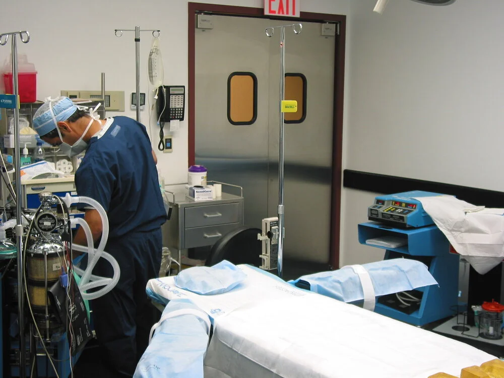 A healthcare professional in blue scrubs, wearing a surgical mask and cap, working in an operating room with medical equipment, IV stand, and a patient bed.