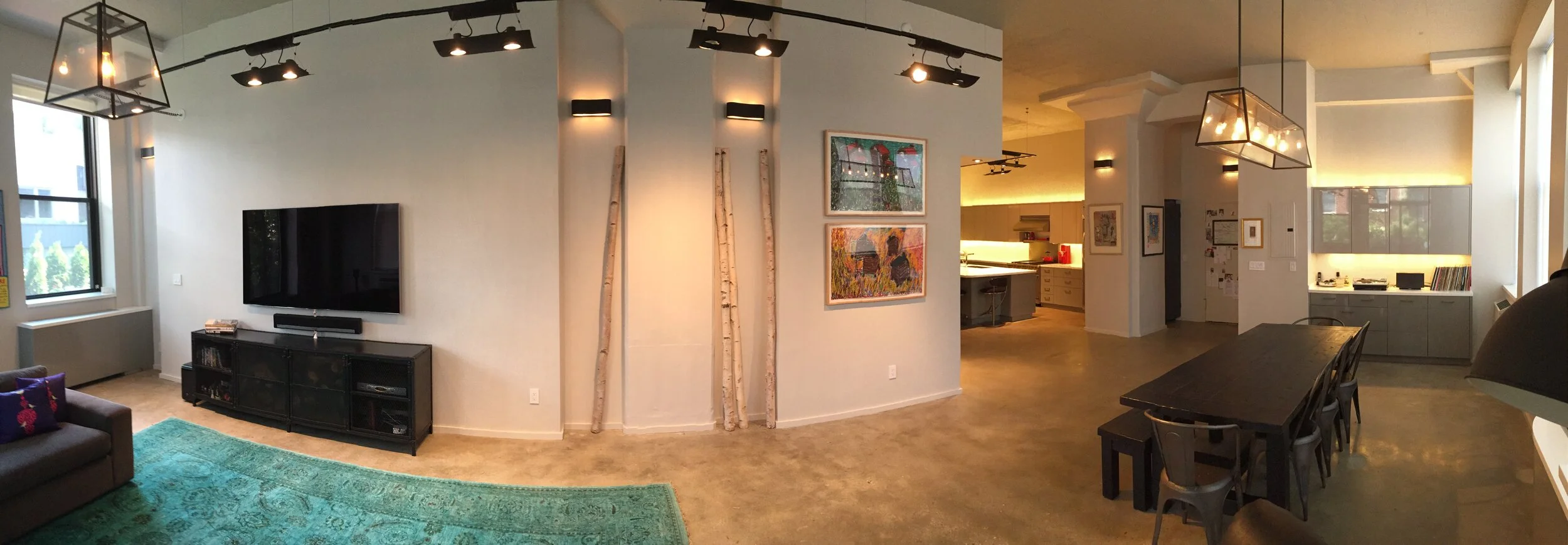 Living room with a wall-mounted flat-screen TV, black entertainment console, a teal rug, a gray sofa with decorative pillows, a window, and a vintage-style light fixture on the ceiling. An artwork featuring colorful abstract landscapes hangs on the w