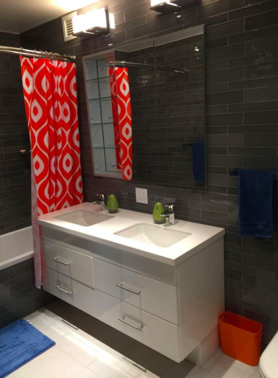 A modern bathroom with a double sink vanity, black tiled walls, a large mirror, a window, a shower with a patterned red and white curtain, and blue and orange accessories.