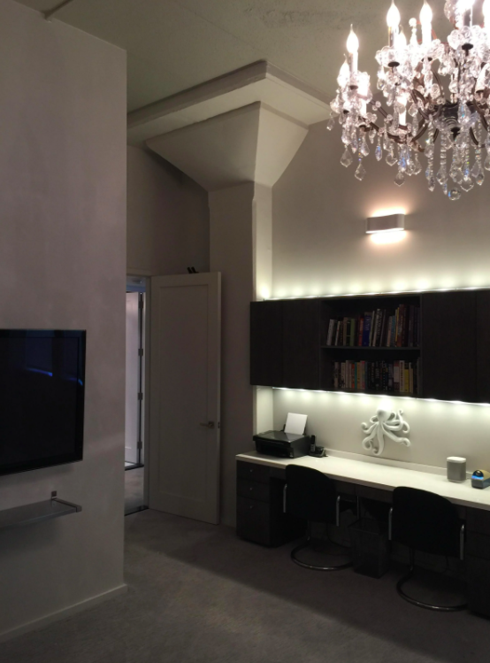 Interior of a modern living space with a chandelier, wall-mounted bookshelf with books, a white desk with electronic devices, two chairs, a wall-mounted TV, and neutral-colored walls and carpeting.