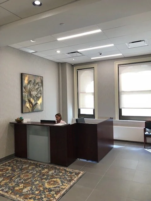 A woman sitting at a reception desk in a modern office with two large windows, a painting on the wall, and a patterned rug.