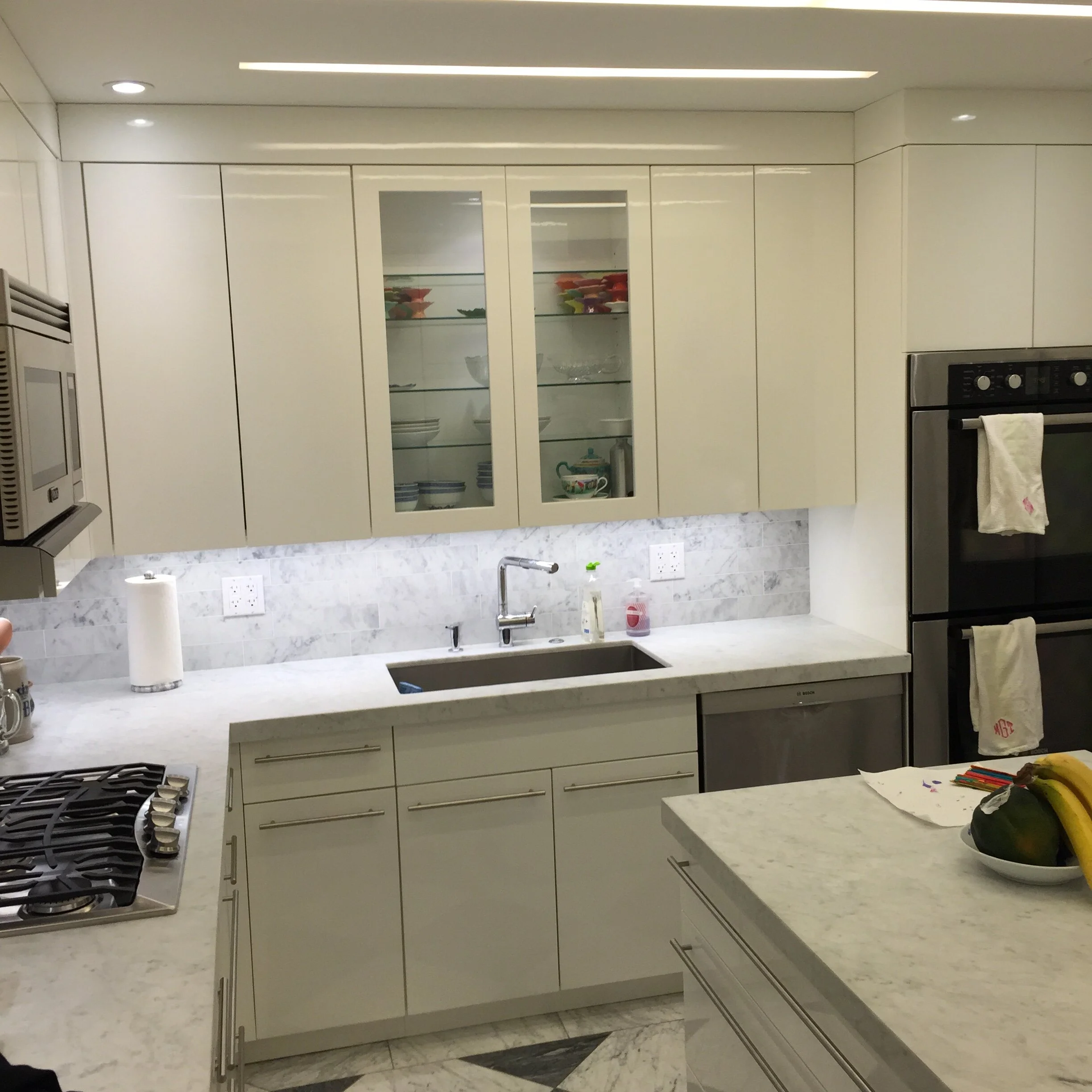 Modern kitchen with white cabinets, marble countertops, and stainless steel appliances, including a stovetop, microwave, and double oven. There are dishes, glasses, and a bowl of fruit on the counters.