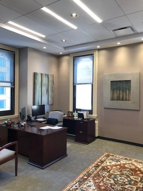 Office with two large windows, a wooden desk with a computer and office supplies, a chair, and abstract artwork on the walls. There is also a patterned rug on the floor.