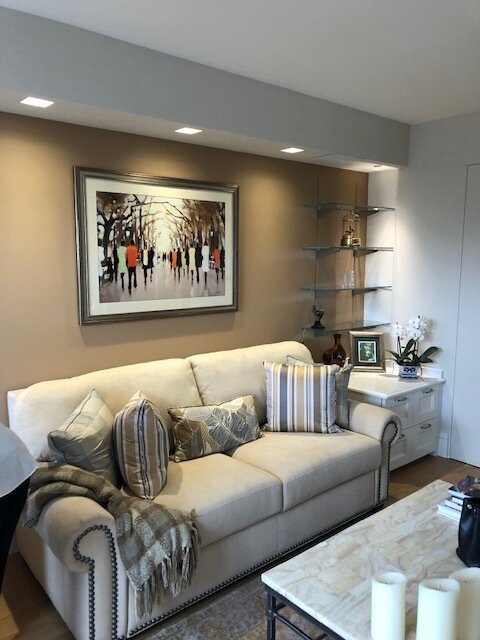 Living room with a white sofa adorned with striped and patterned pillows, a beige throw blanket, a white side table with a potted plant and framed photo, glass shelves with decorative items, a framed painting of people walking in a park, and a light 